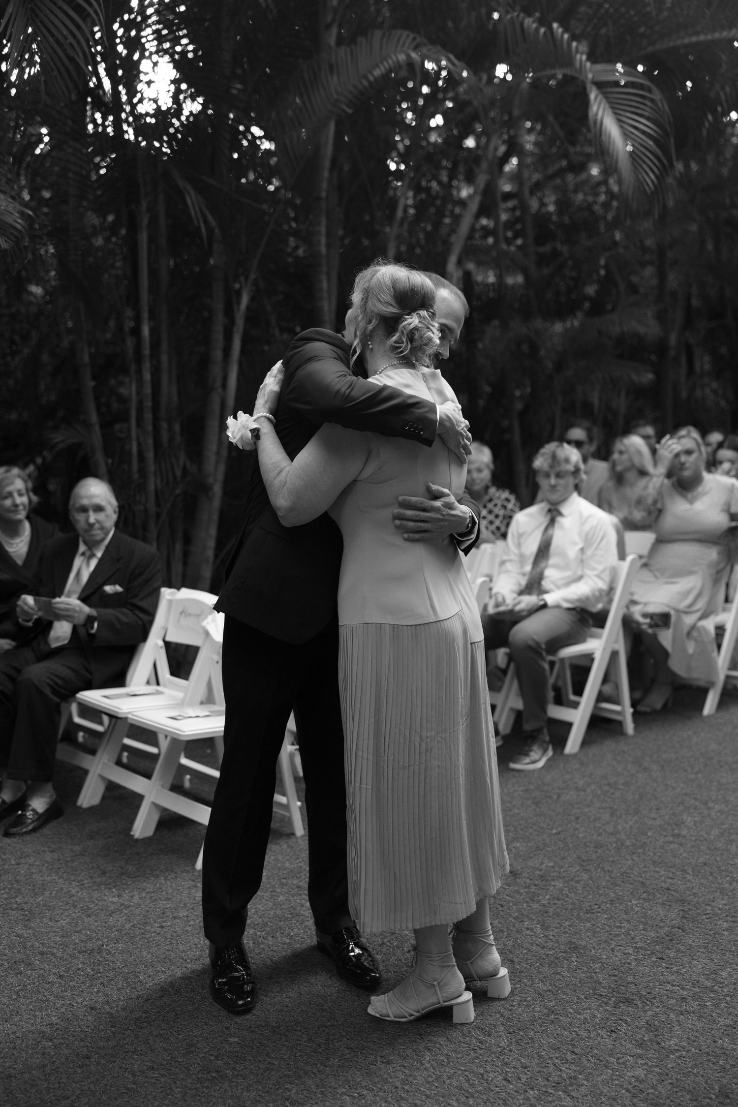 Emotional moment at outdoor wedding ceremony with guests seated on white chairs in garden setting.