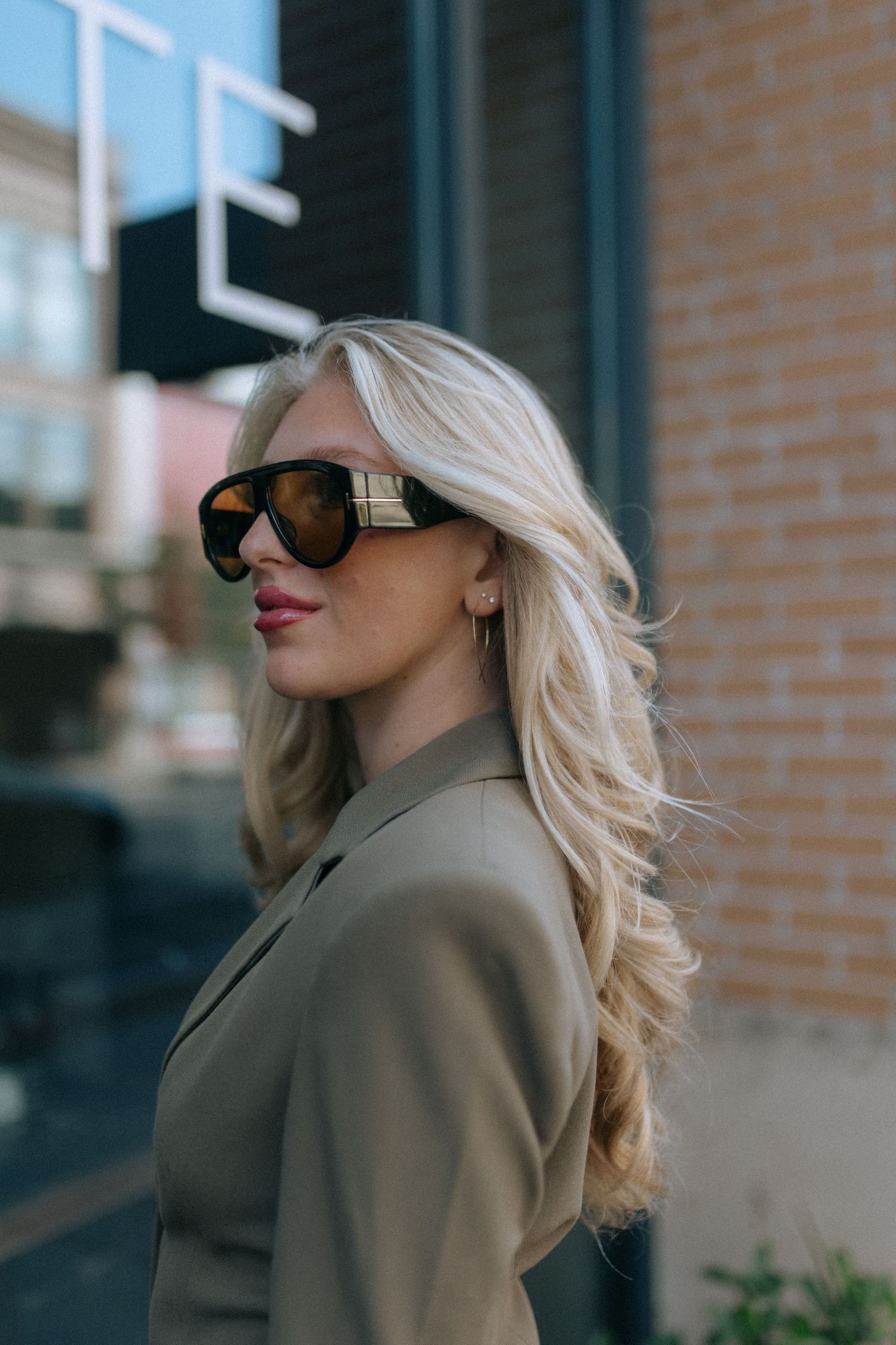 Woman with blonde hair wearing sunglasses and tan blazer poses against brick wall.