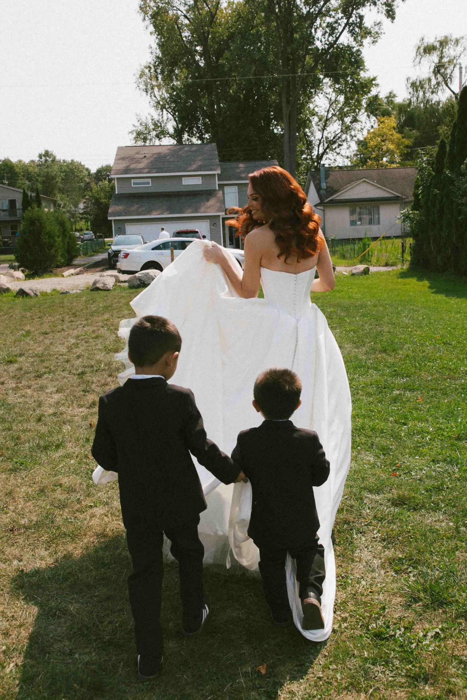 Two small children in formal attire follow someone in a flowing white dress across a lawn.