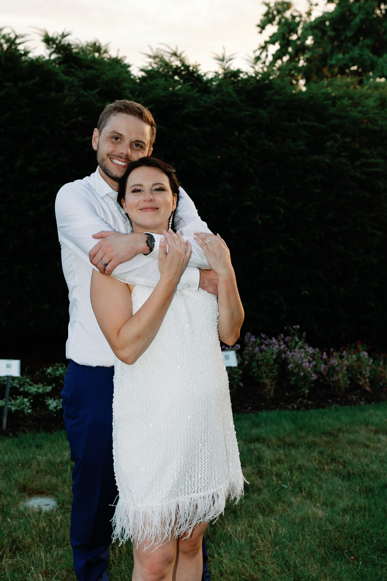 Intimate moment captured between couple in white dress and navy suit on garden grounds.