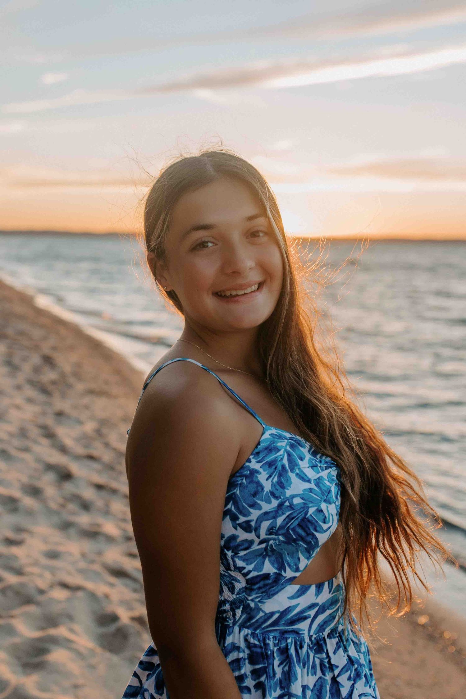 Person in blue floral dress poses on beach at sunset with hair flowing in ocean breeze.