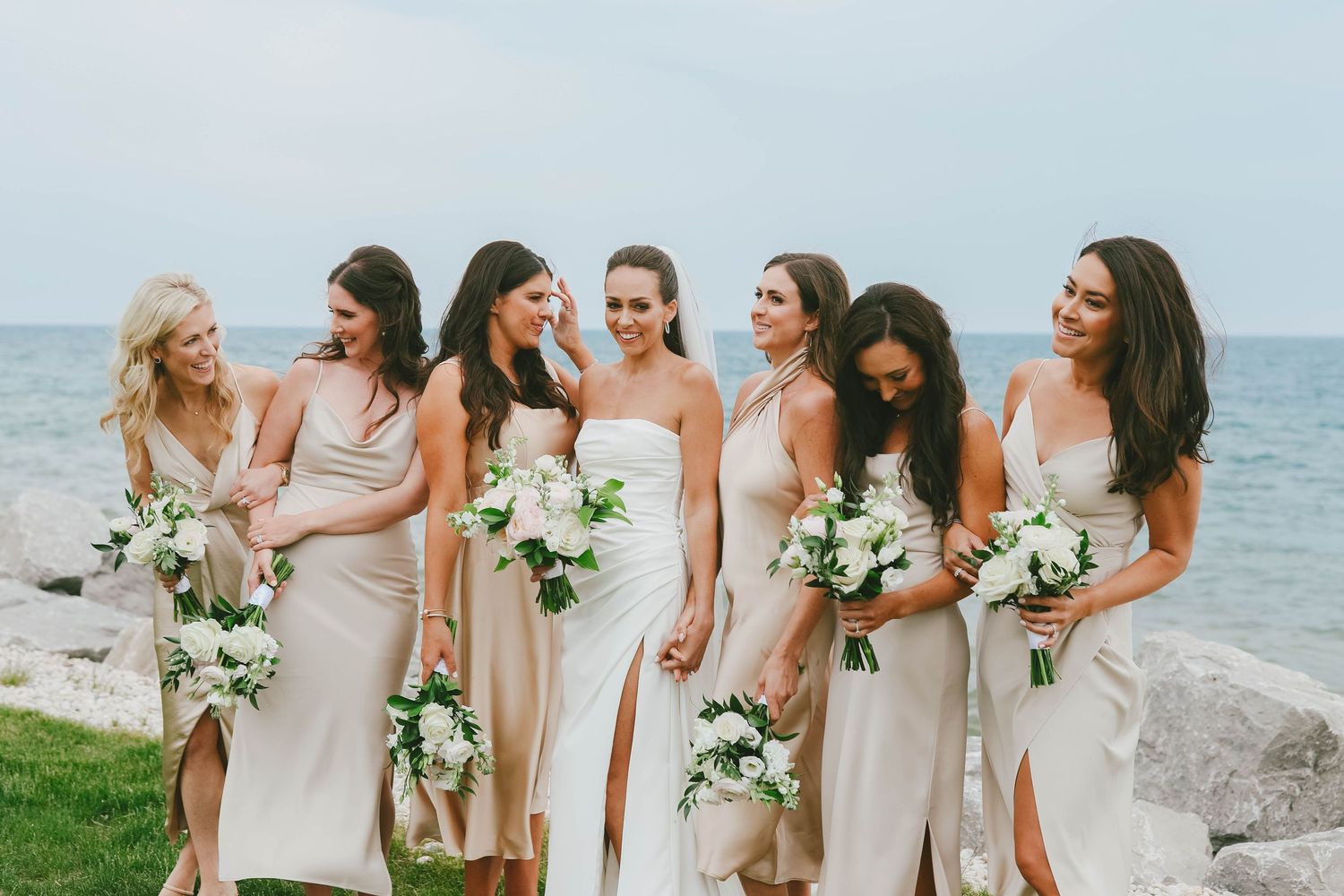 Beachside wedding party in neutral dresses holding white floral bouquets against ocean backdrop.