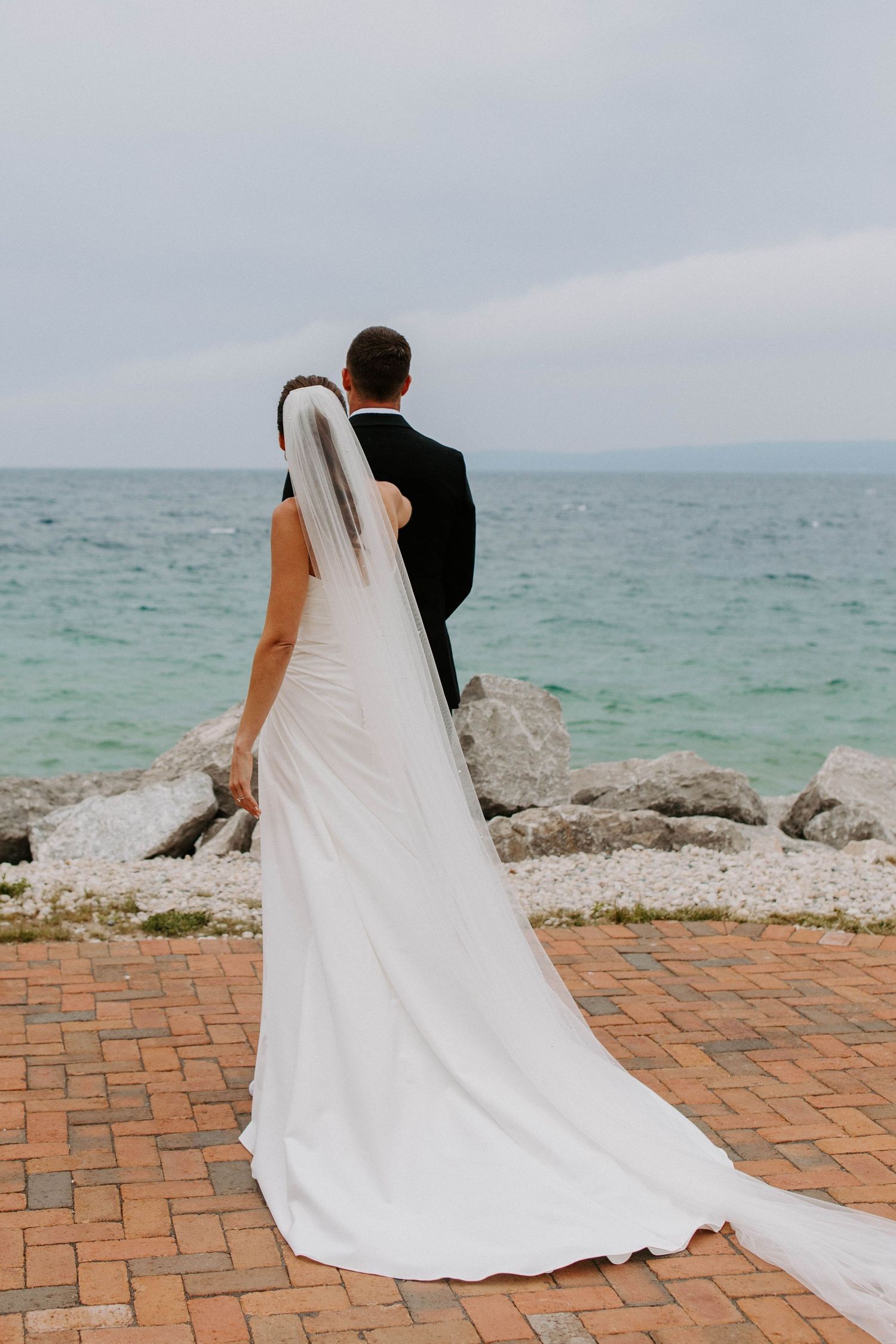 A wedding couple walks along a brick pathway with ocean waves in the background.