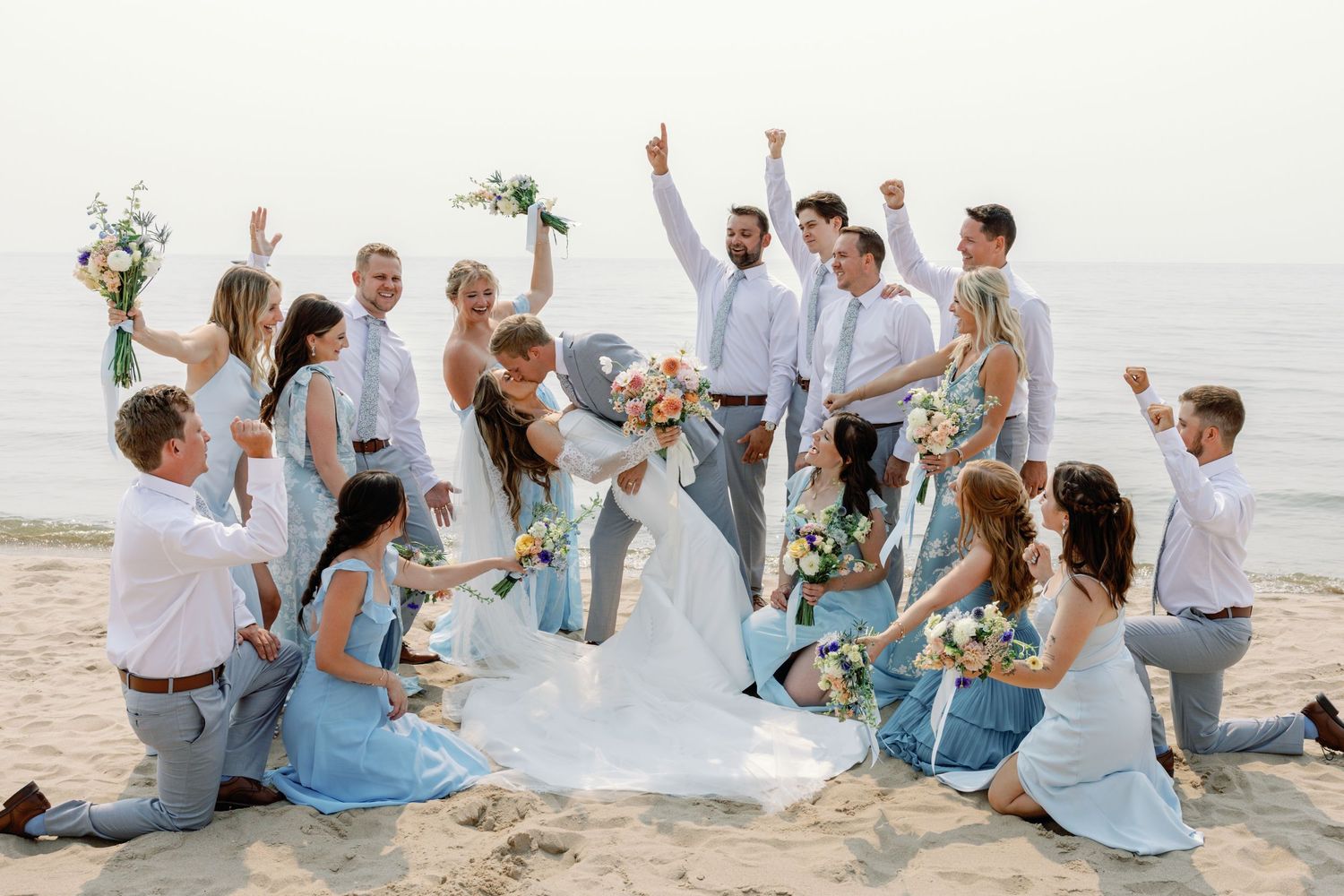Wedding party celebrates joyfully on a sandy beach with bridesmaids in light blue dresses.