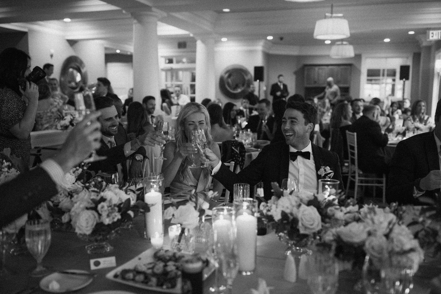 Black and white candid of wedding guests enjoying dinner at tables decorated with flowers and candles.