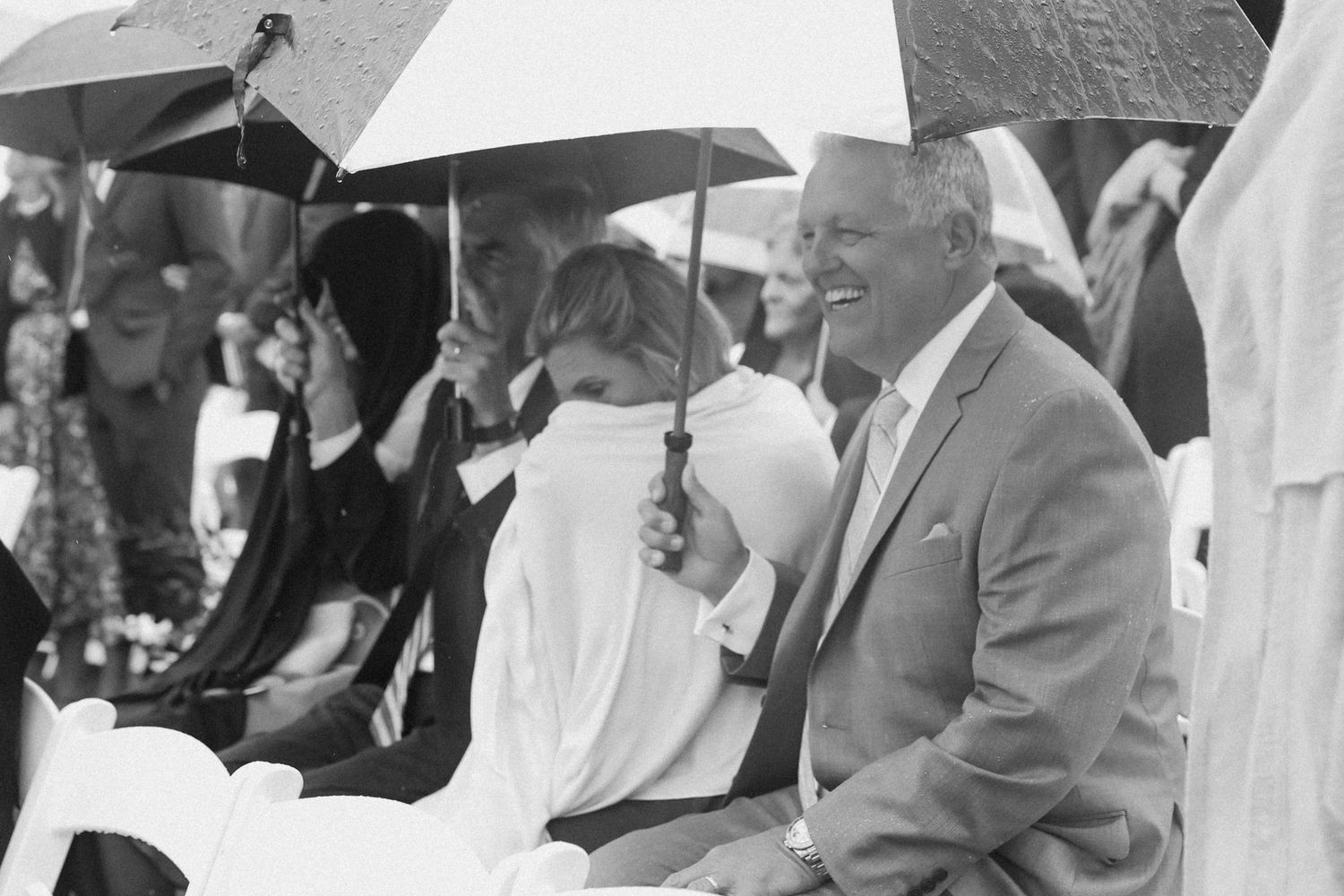 Black and white photo of wedding guests holding umbrellas during an outdoor ceremony.