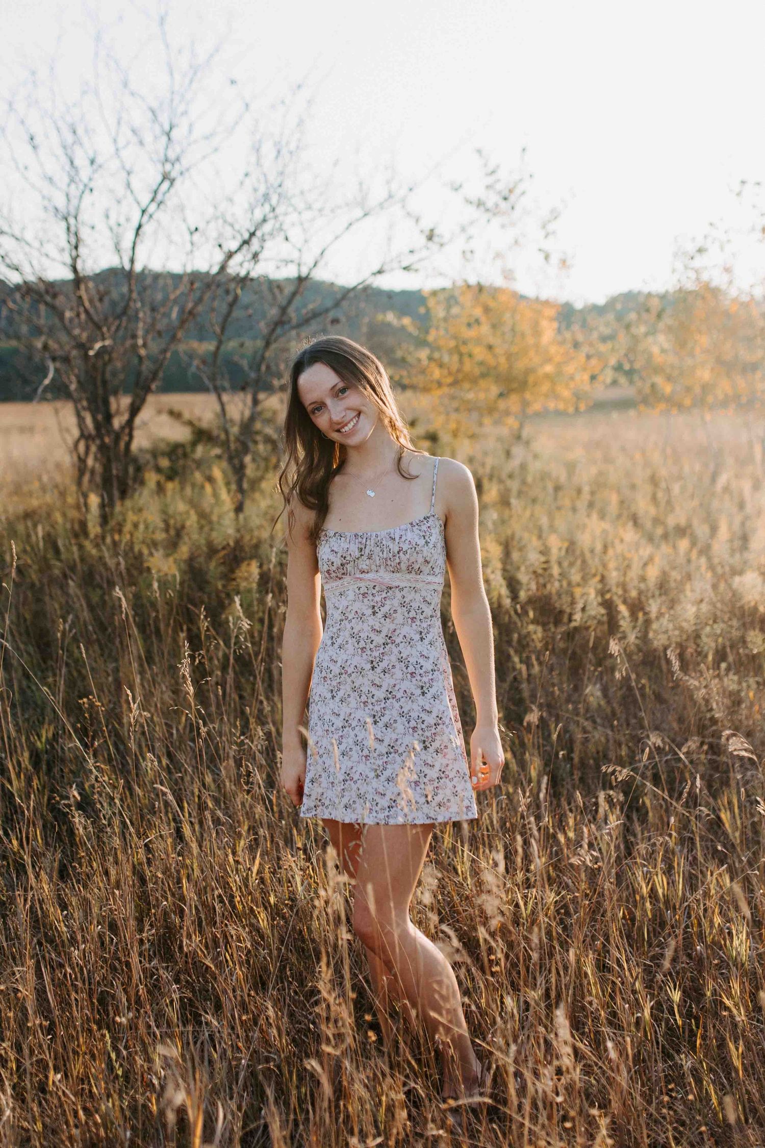 Casual pose in a floral dress amidst wild grasses during sunset.