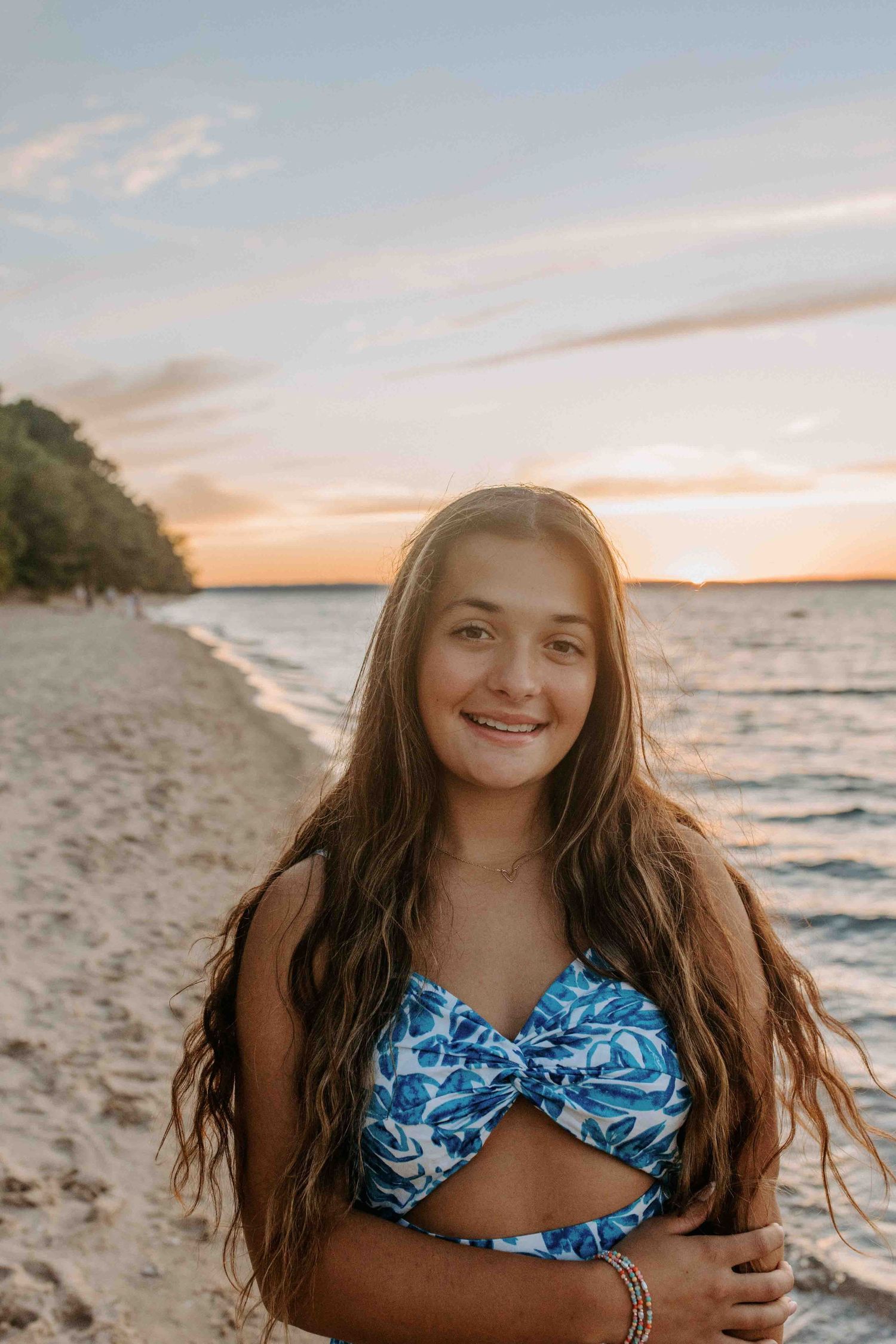 Person wearing a blue cutout dress smiles while standing on a beach at sunset.