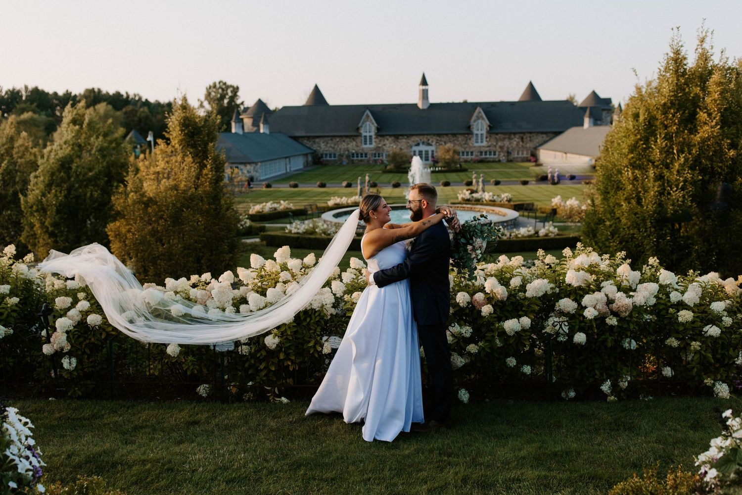 Wedding party poses in golden sunlight among white flowers in a garden setting.