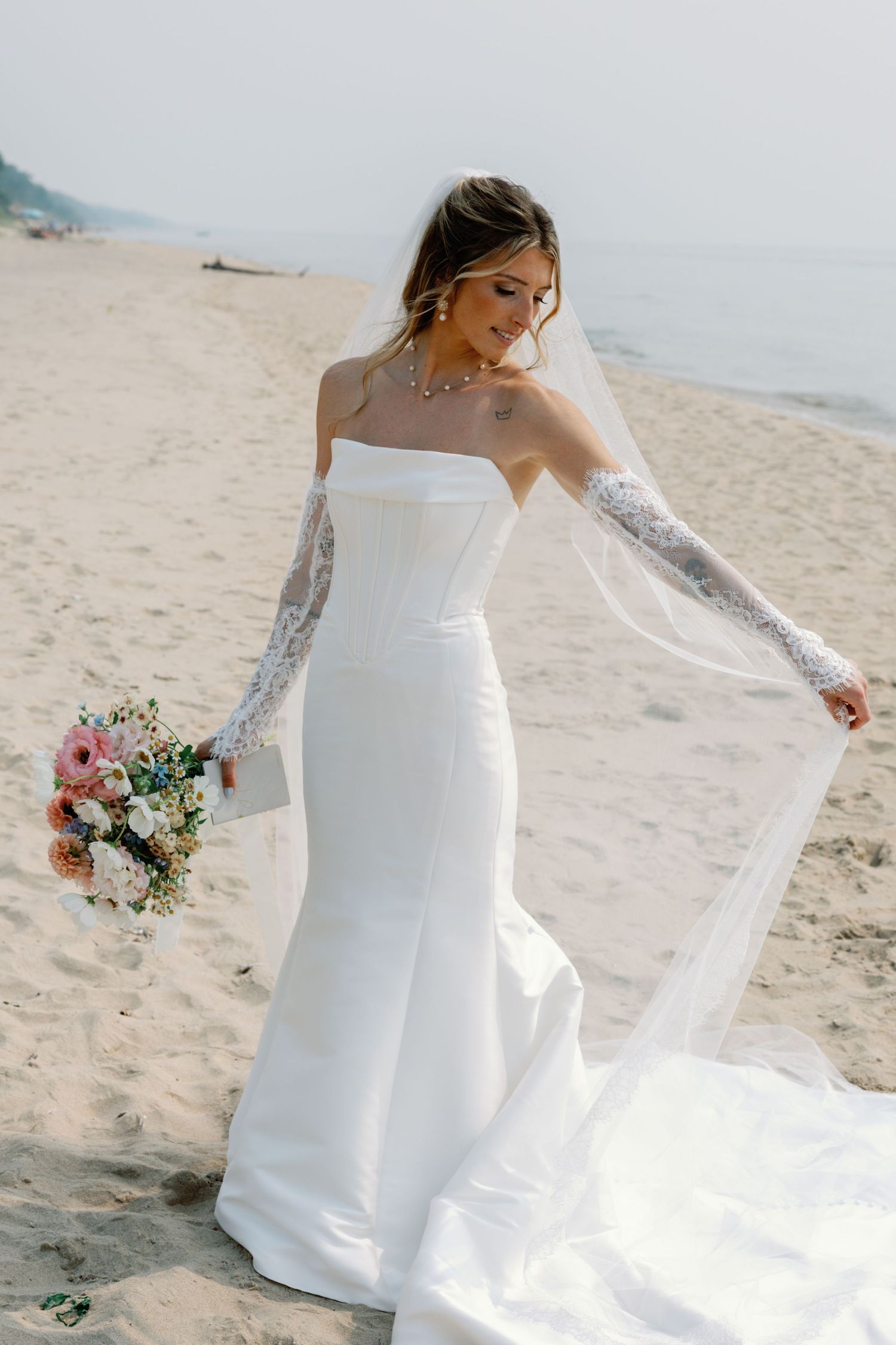 A flowing white wedding dress with lace sleeves billows in the ocean breeze on a sandy beach.