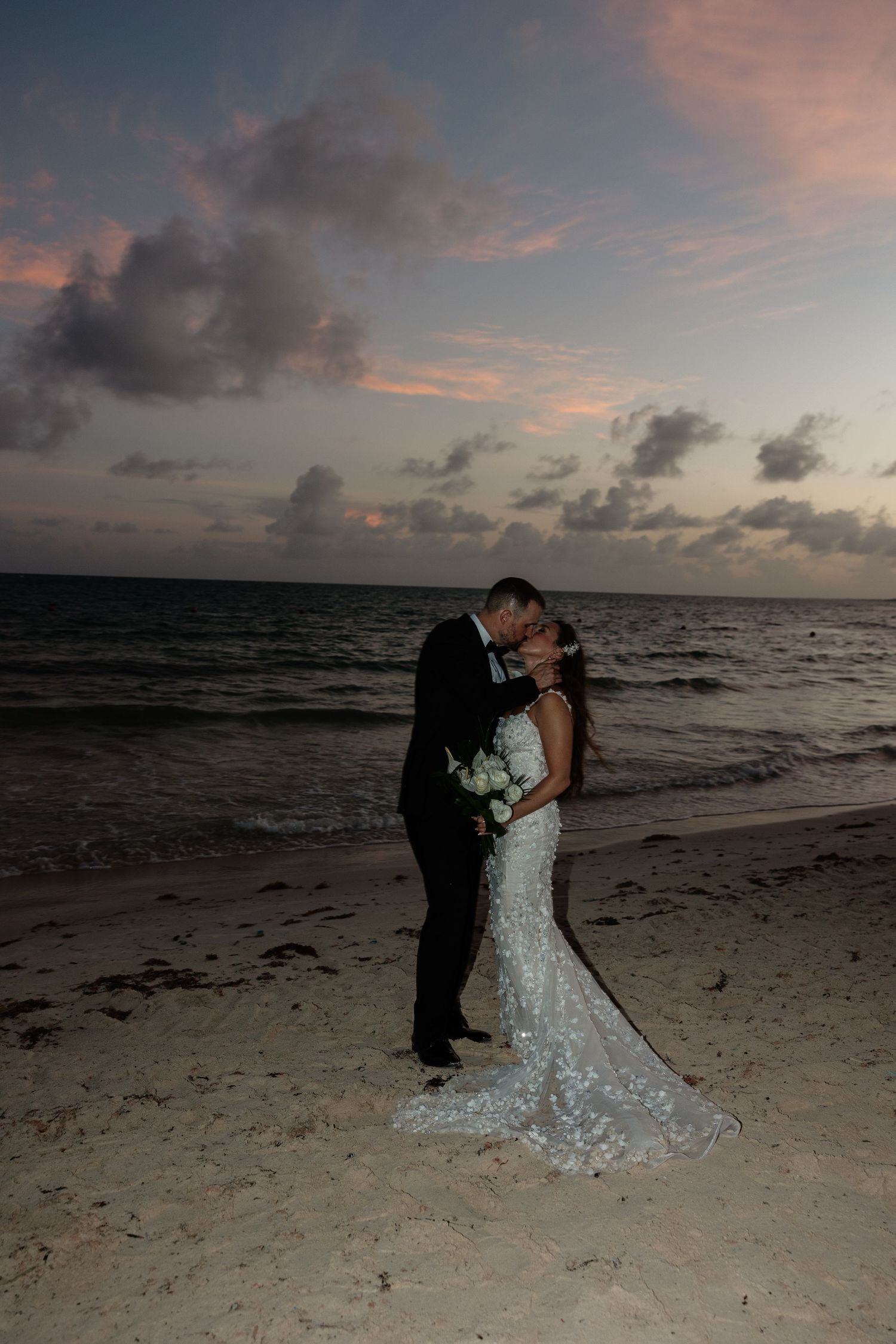 Newlyweds share an intimate moment on the beach at sunset with dramatic clouds and ocean views.