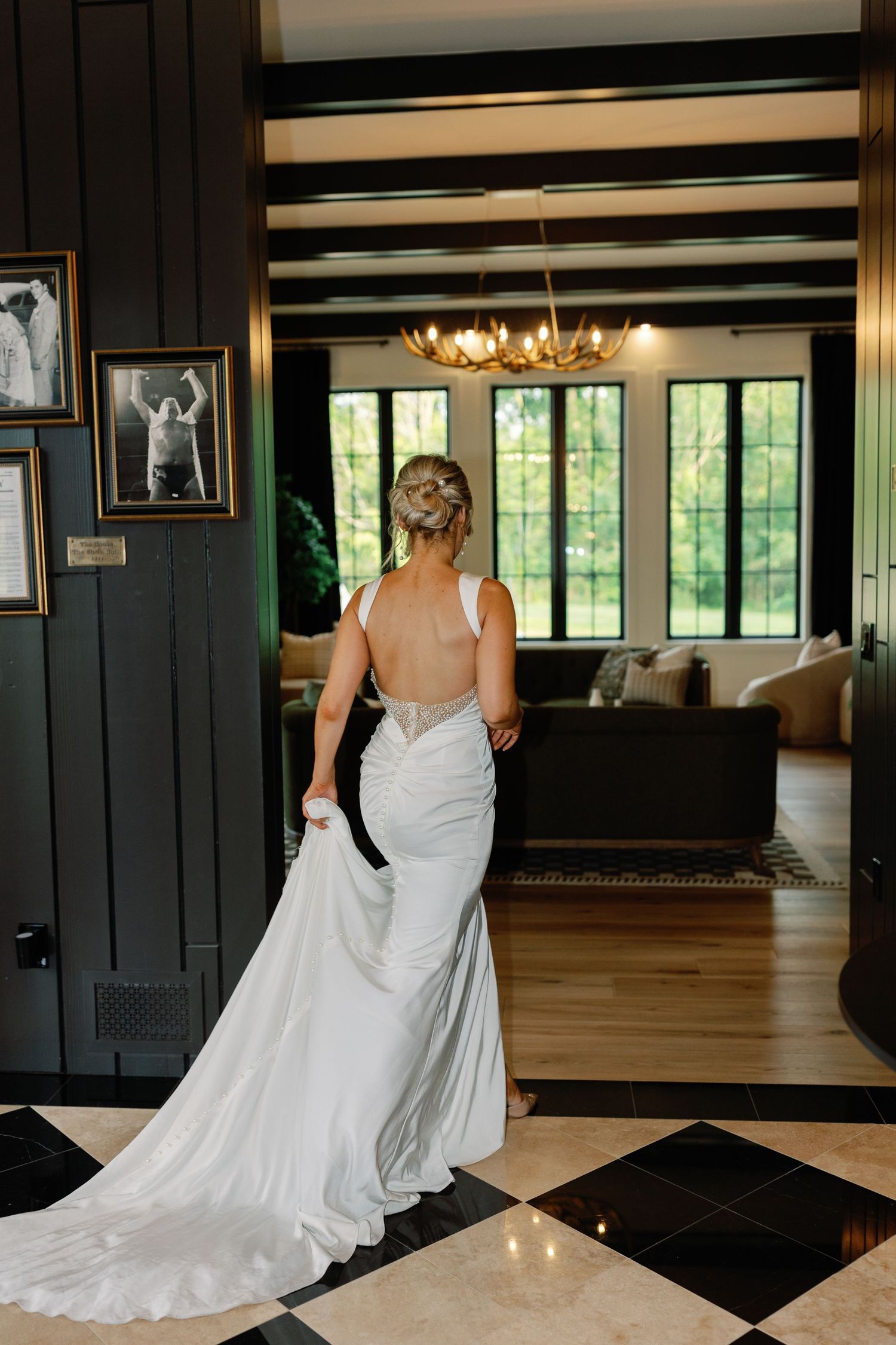 A bride in a flowing white wedding dress walks across a black and white checkered ballroom floor with windows and chandeliers.