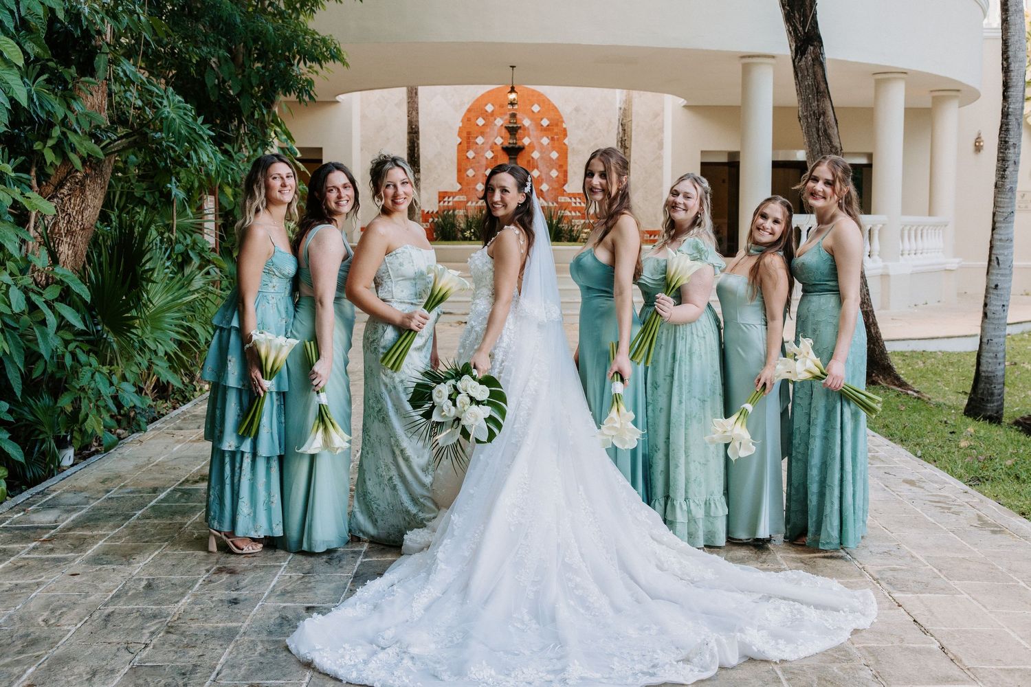 Bridesmaids in seafoam green dresses pose with bride in elegant white gown against stone building.