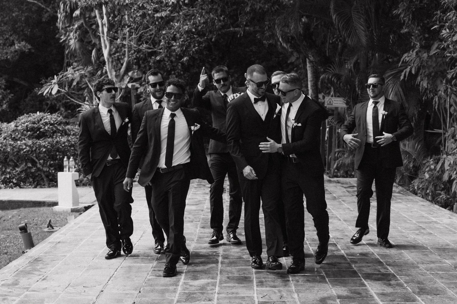 Wedding party members in dark suits leaping in unison on stone steps surrounded by greenery.