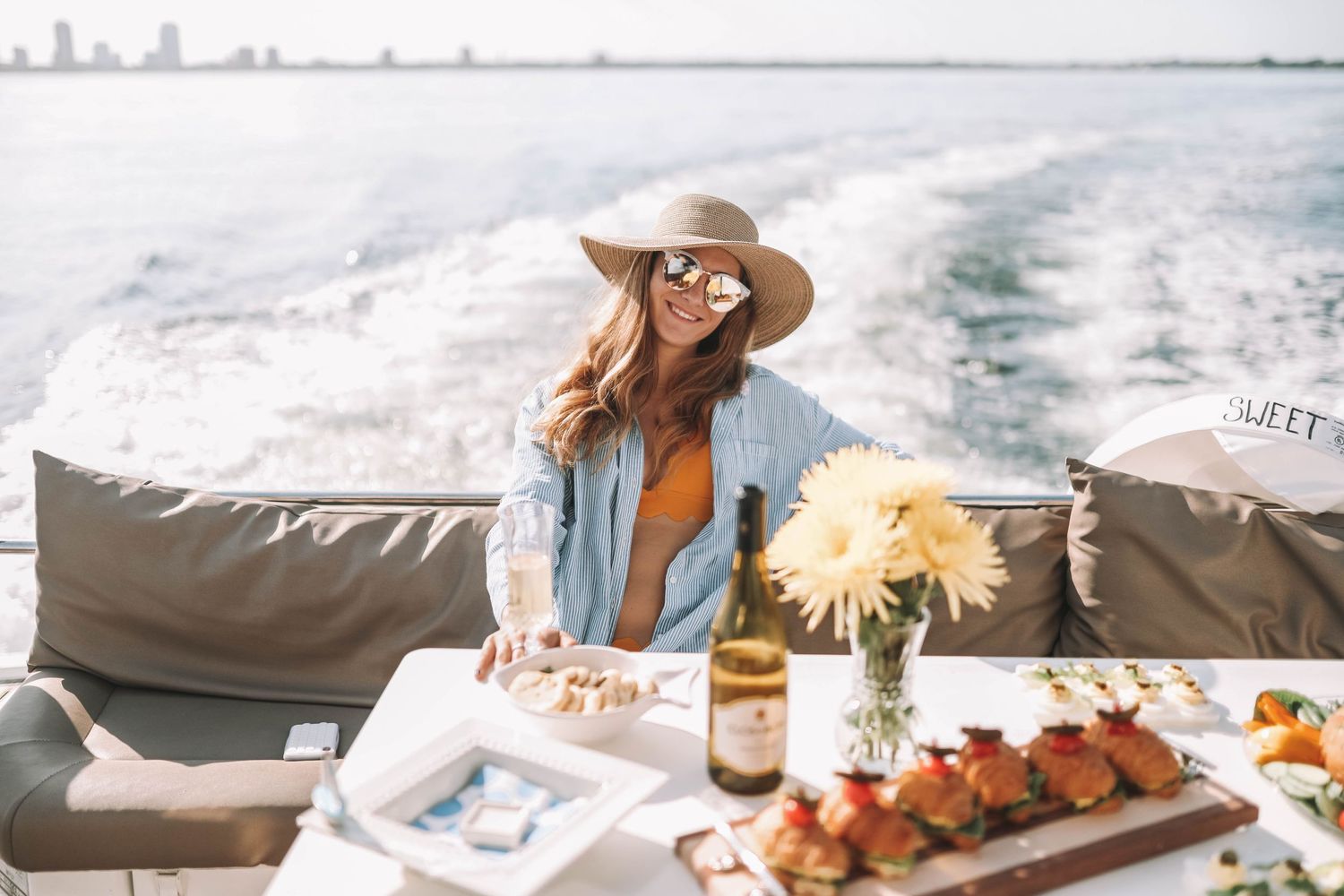 Luxury yacht experience featuring champagne, appetizers, and sun hats on a white cushioned deck by the ocean.