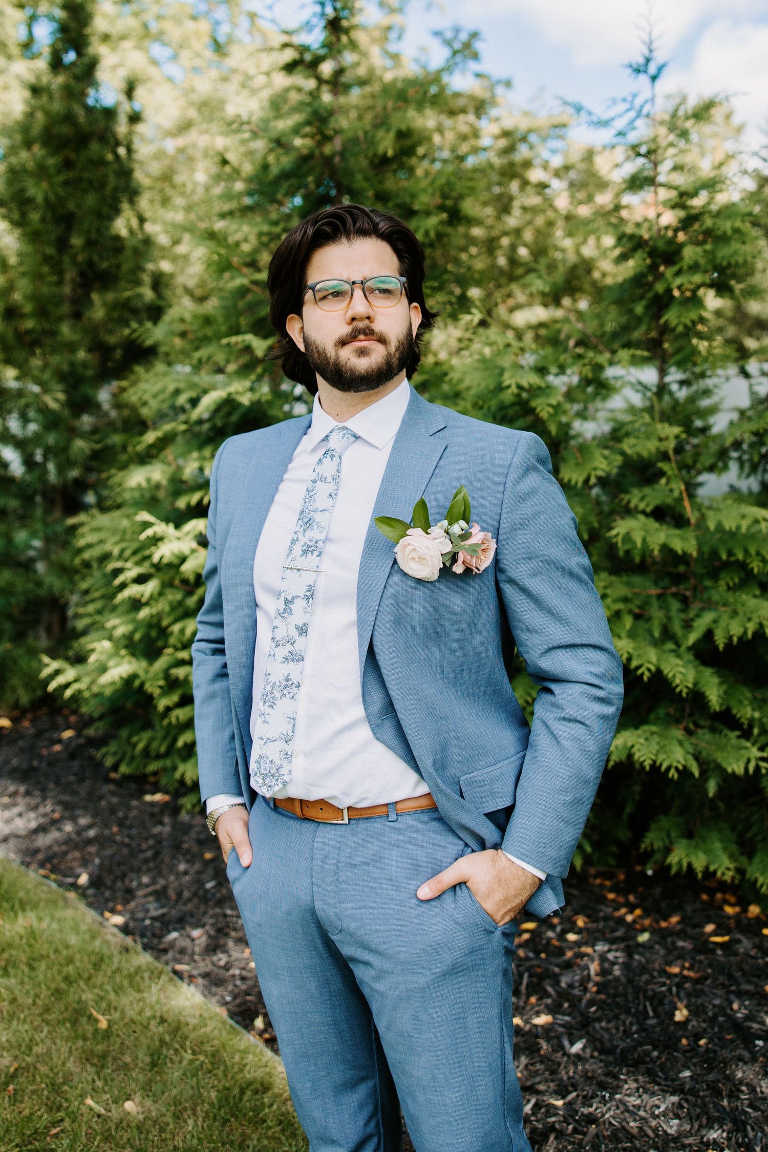 Wedding attire featuring a light blue suit with floral tie against a green outdoor backdrop.