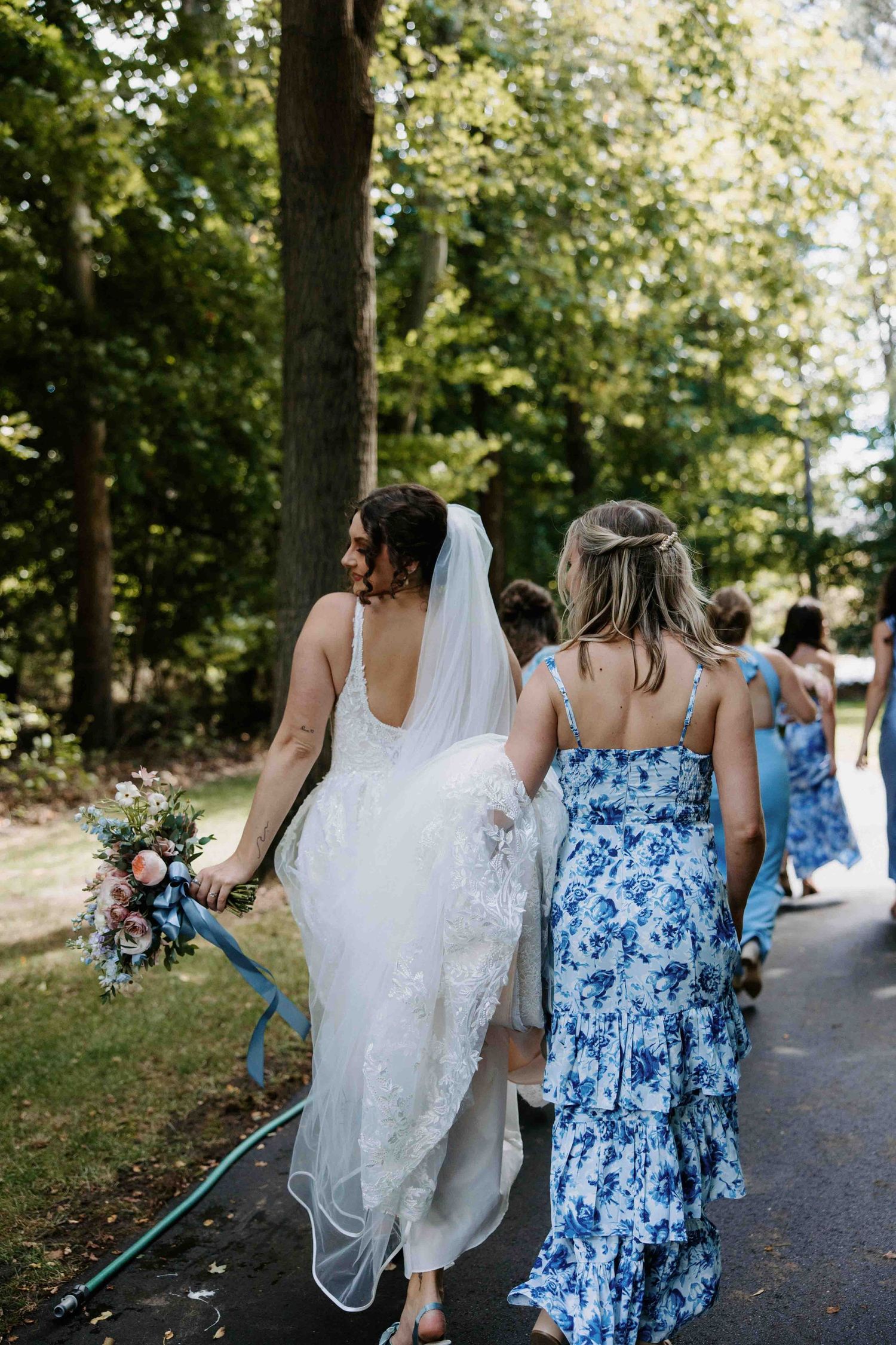 Bridesmaids in blue floral and solid dresses walk with the bride along a shaded path.