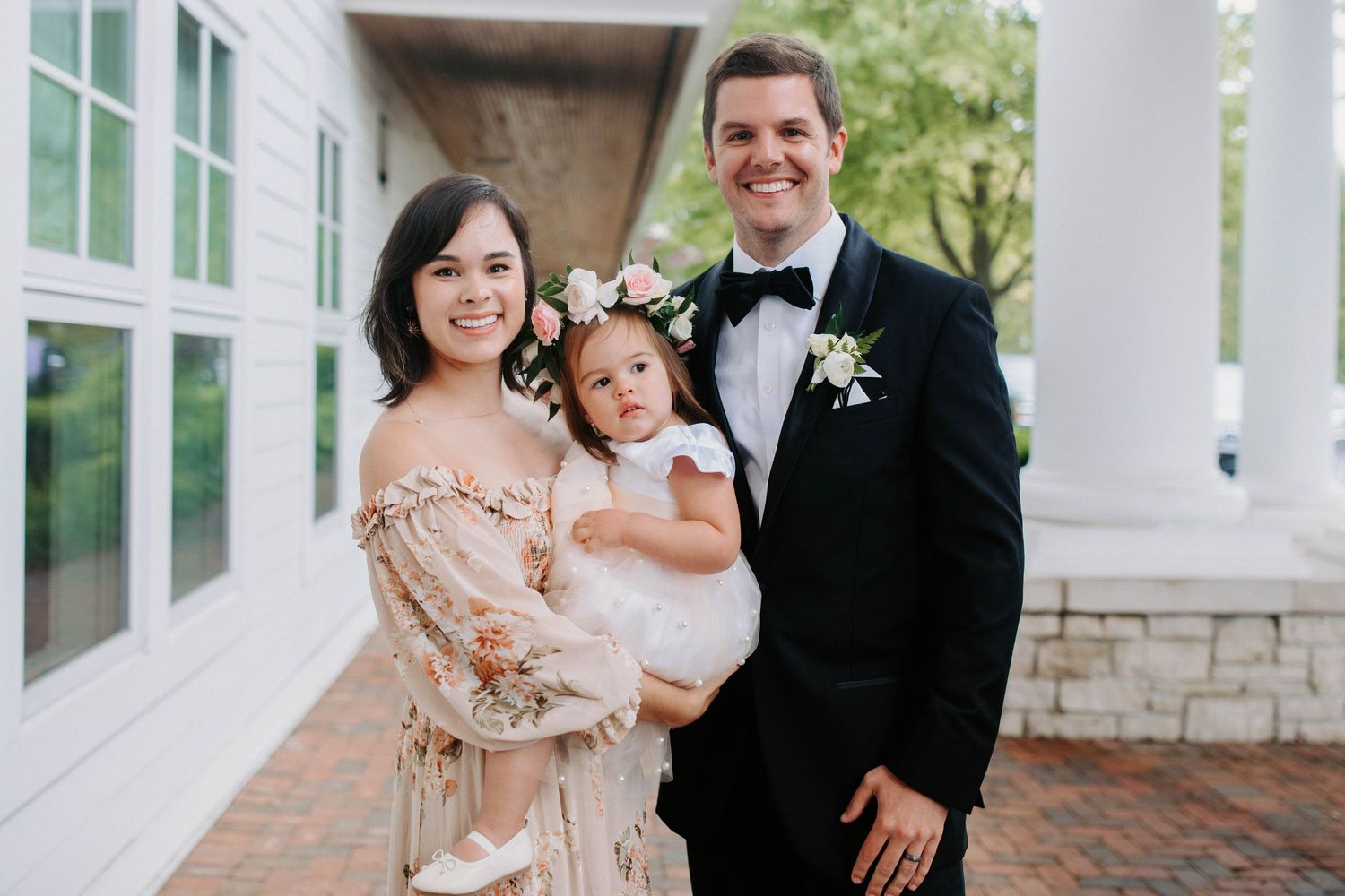 A couple and child pose together on a white columned porch during a formal event.
