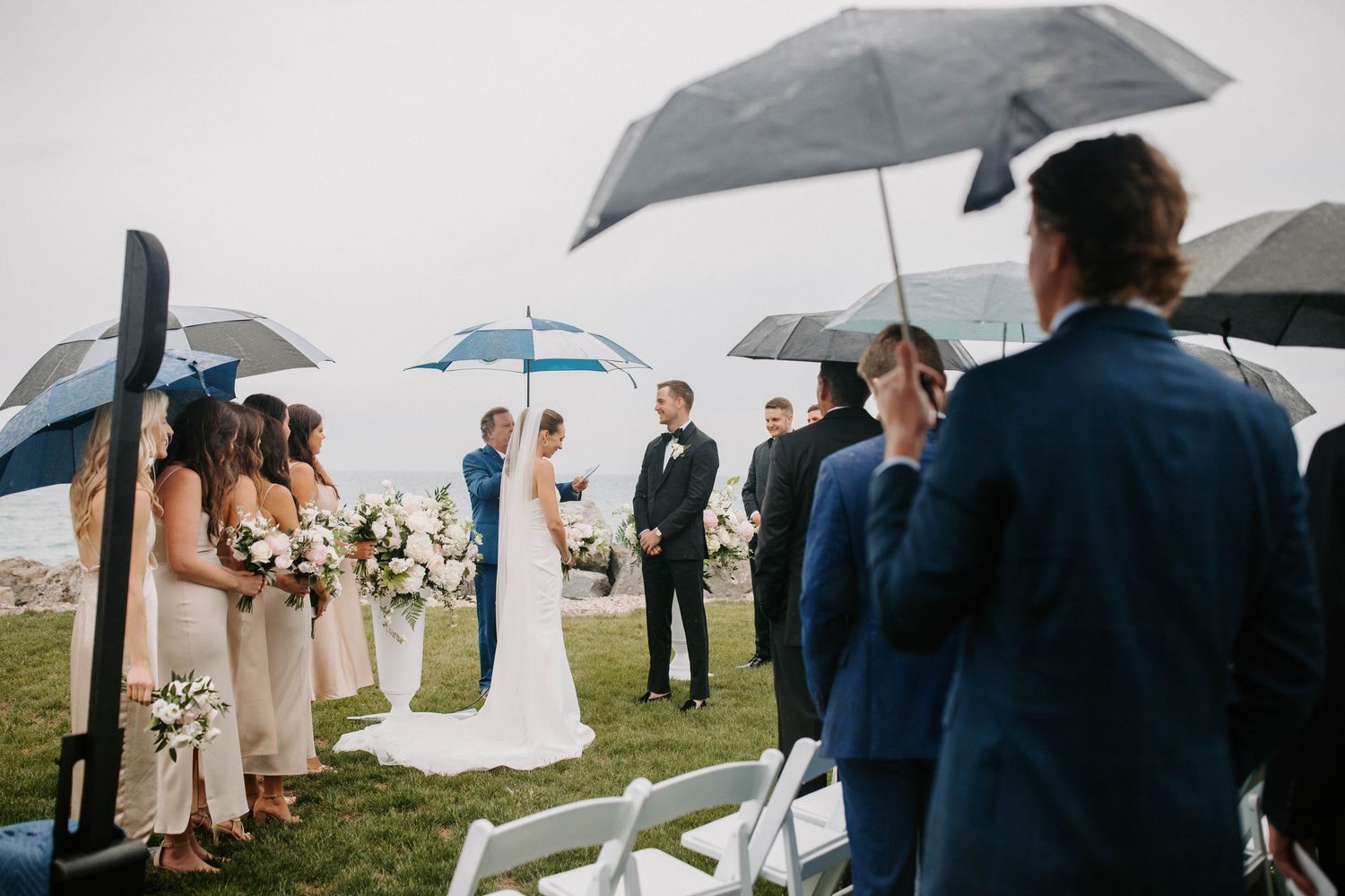 Wedding guests and party members gather under umbrellas during an outdoor ceremony.