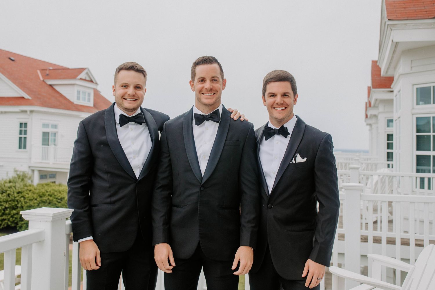 Groomsmen in black tuxedos posing together on oceanfront property.