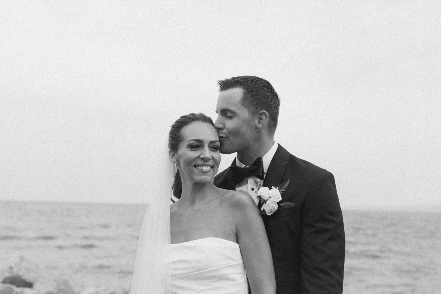 A black and white seaside wedding portrait showing a tender moment between newlyweds.