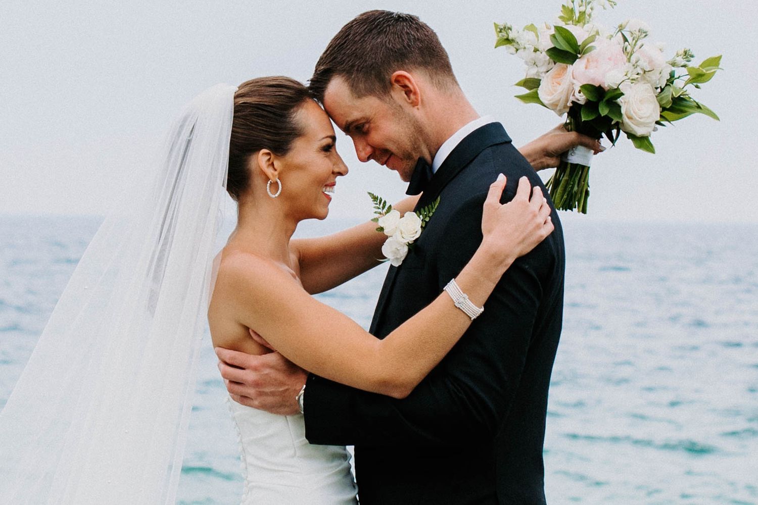 Intimate wedding moment captured against ocean backdrop with flowing veil.