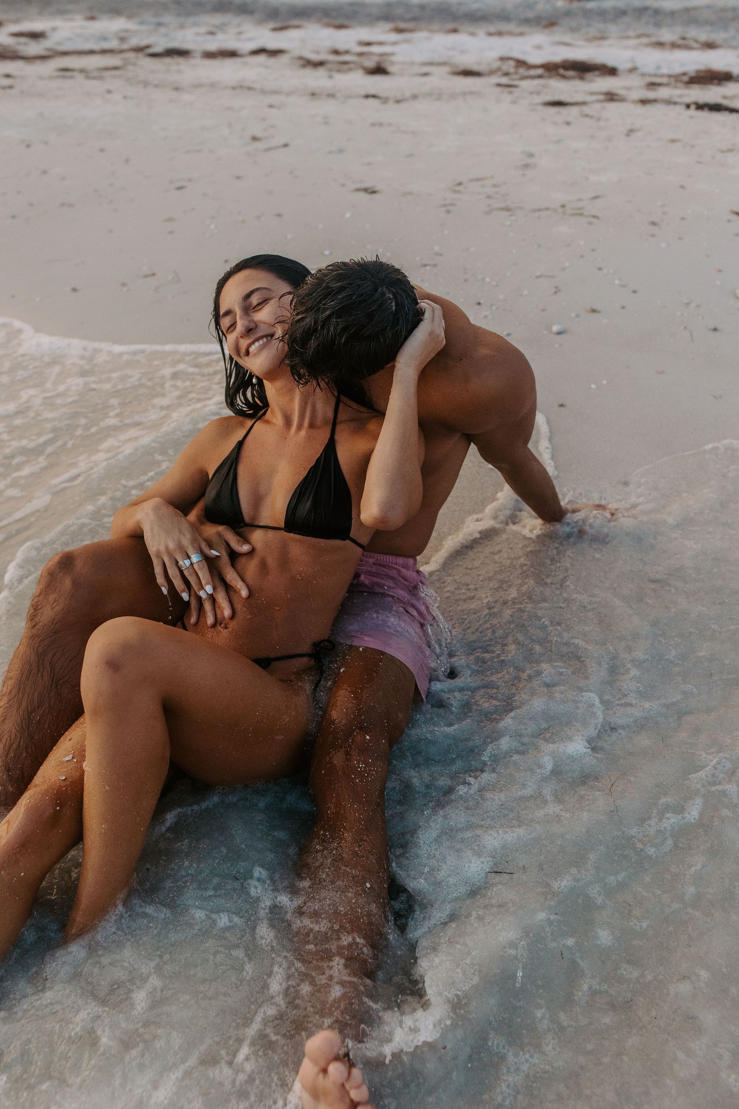 A couple shares romantic moments while lying in shallow beach waters at sunset.