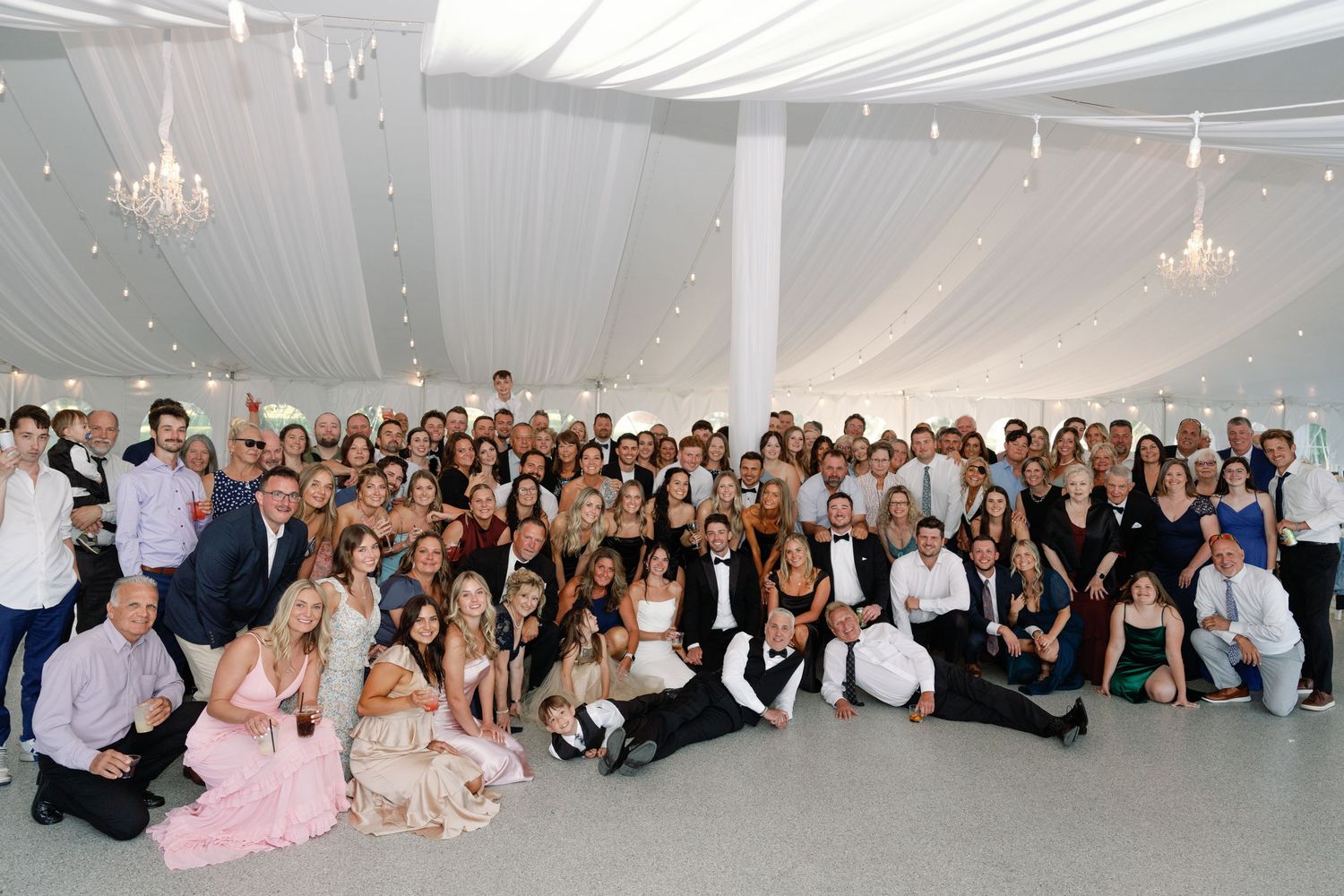 Large group photo of wedding guests gathered together in an elegant indoor venue with chandeliers.