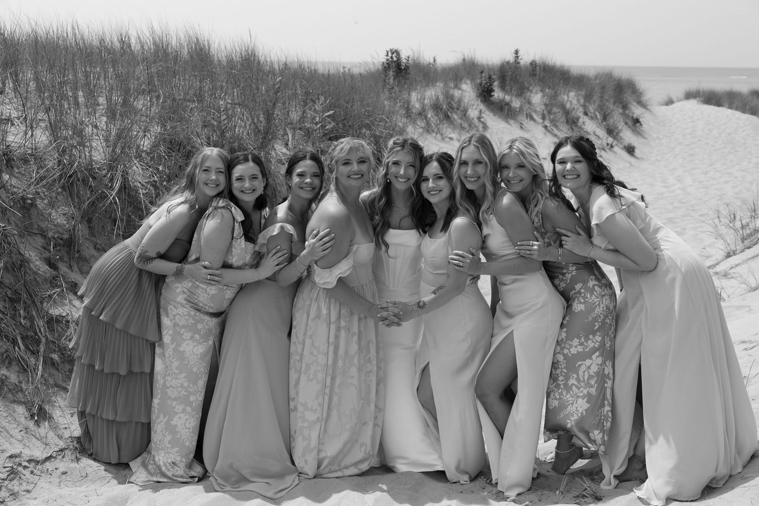 A bridal party in matching dresses poses together on a sandy beach landscape.