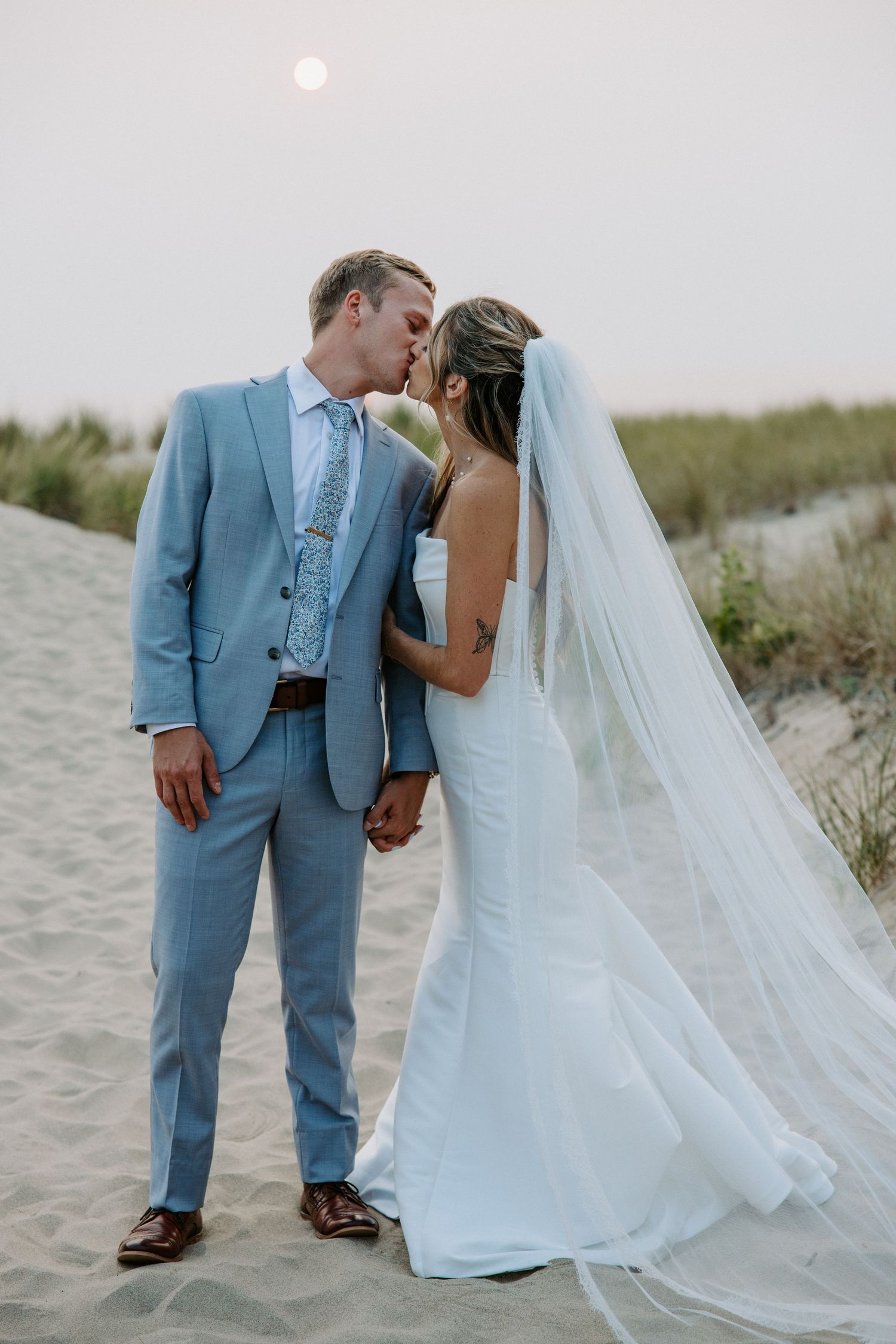 Wedding couple shares intimate moments on sandy beach with flowing veil and light blue suit.