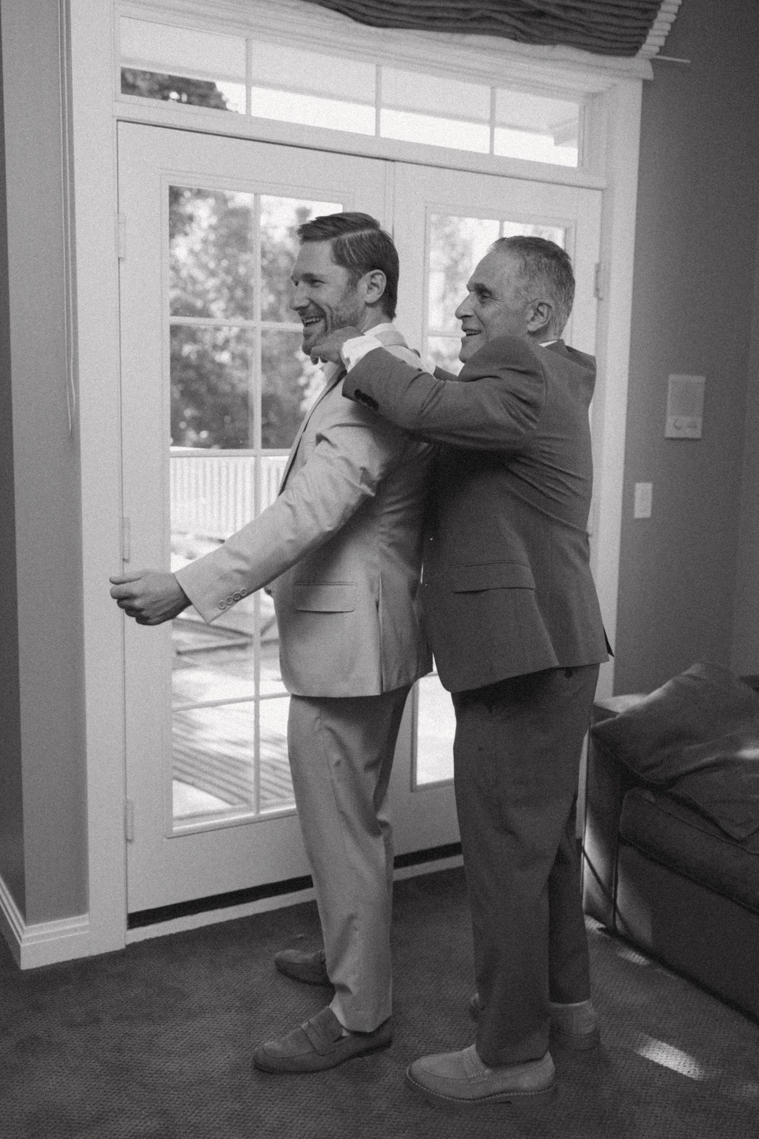 Black and white photo of groomsmen helping each other prepare in a sunlit room.