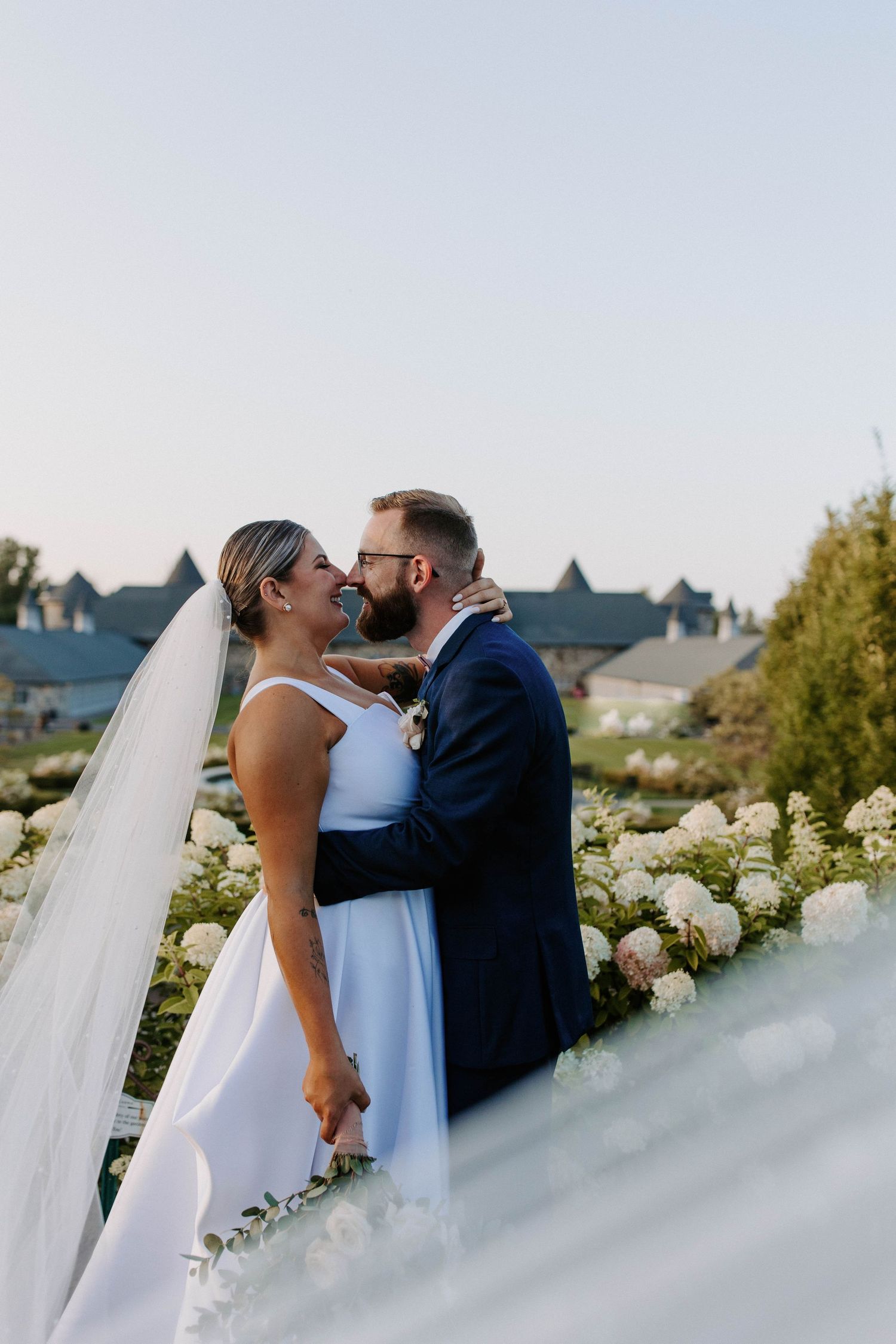 Intimate wedding moment captured through flowing white veil with garden flowers in background.