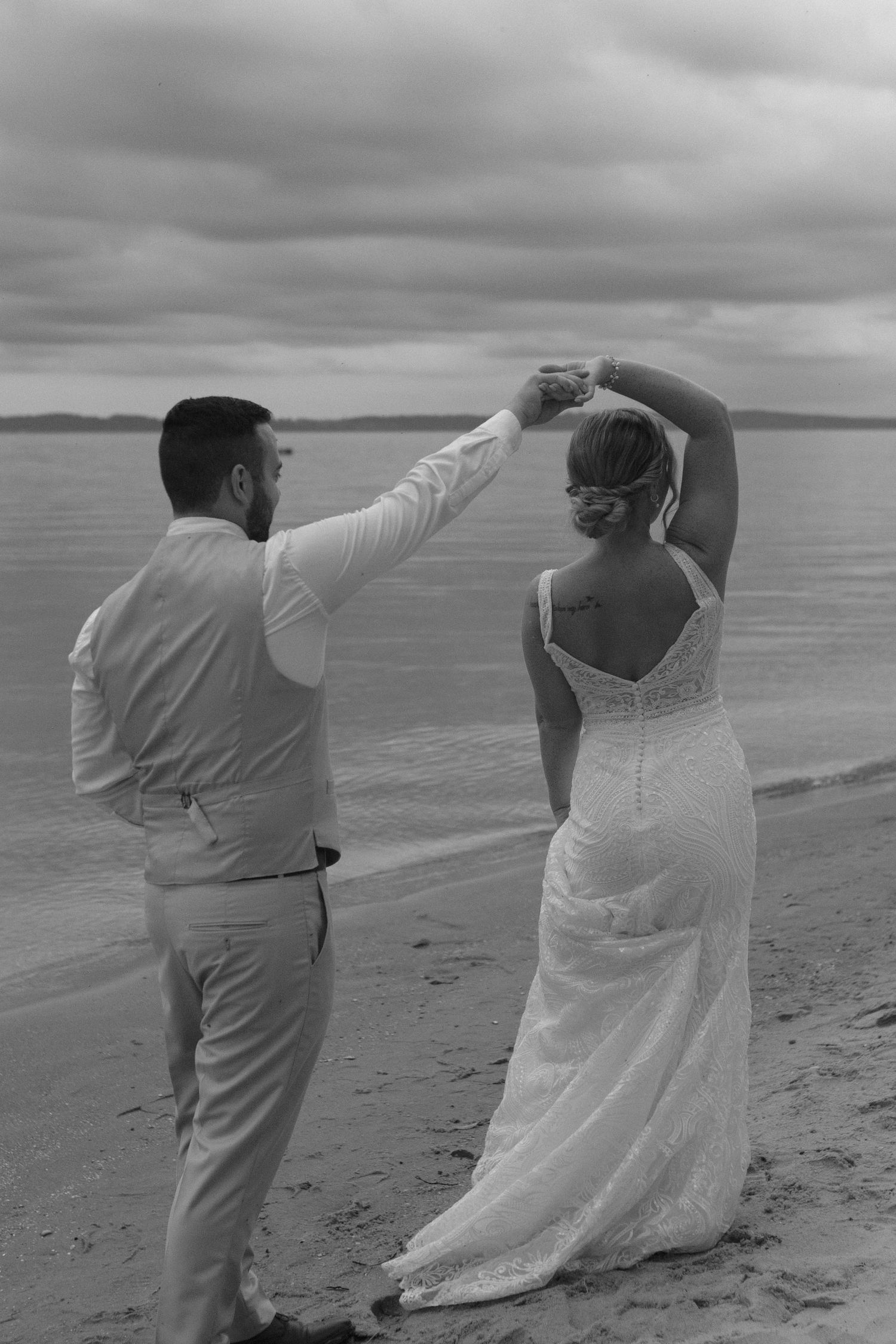 Playful couple in wedding attire strike dramatic poses on a cloudy beach.