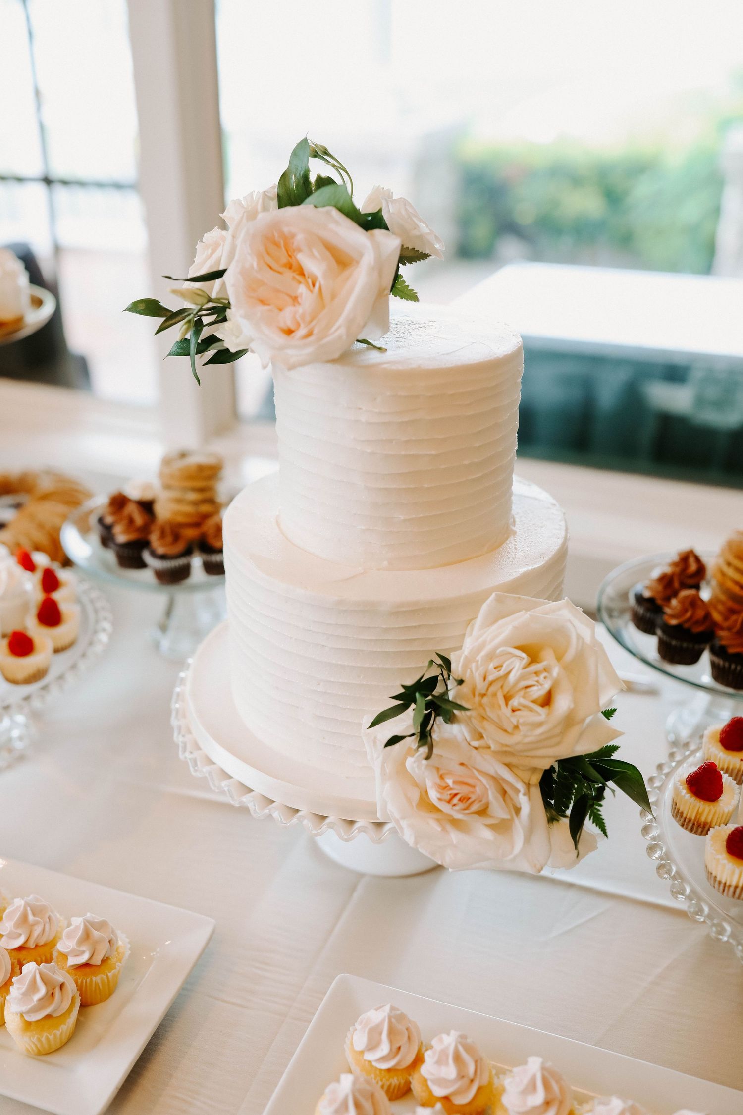 White wedding cakes decorated with fresh roses and surrounded by cupcakes on crystal stands.