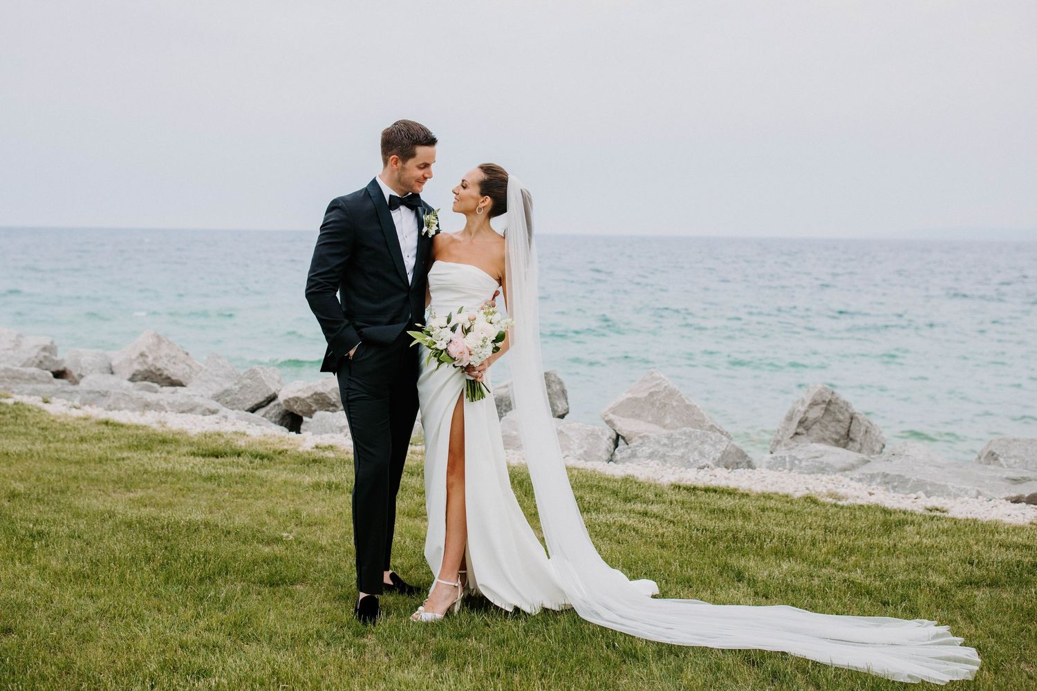 Elegant coastal wedding photoshoot with couple embracing on a scenic oceanfront lawn.