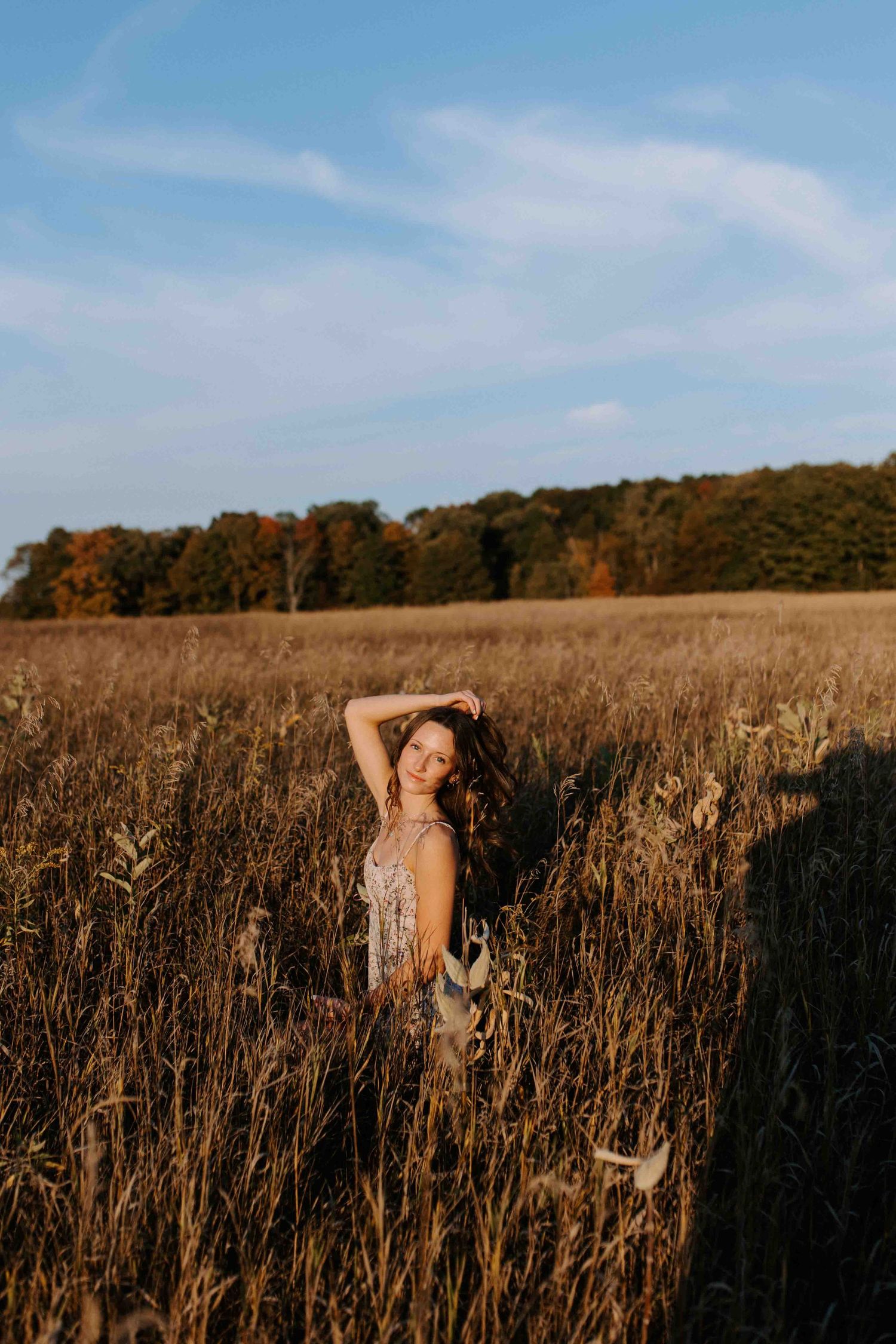 Someone poses gracefully in a field of tall grass during sunset with trees on the horizon.