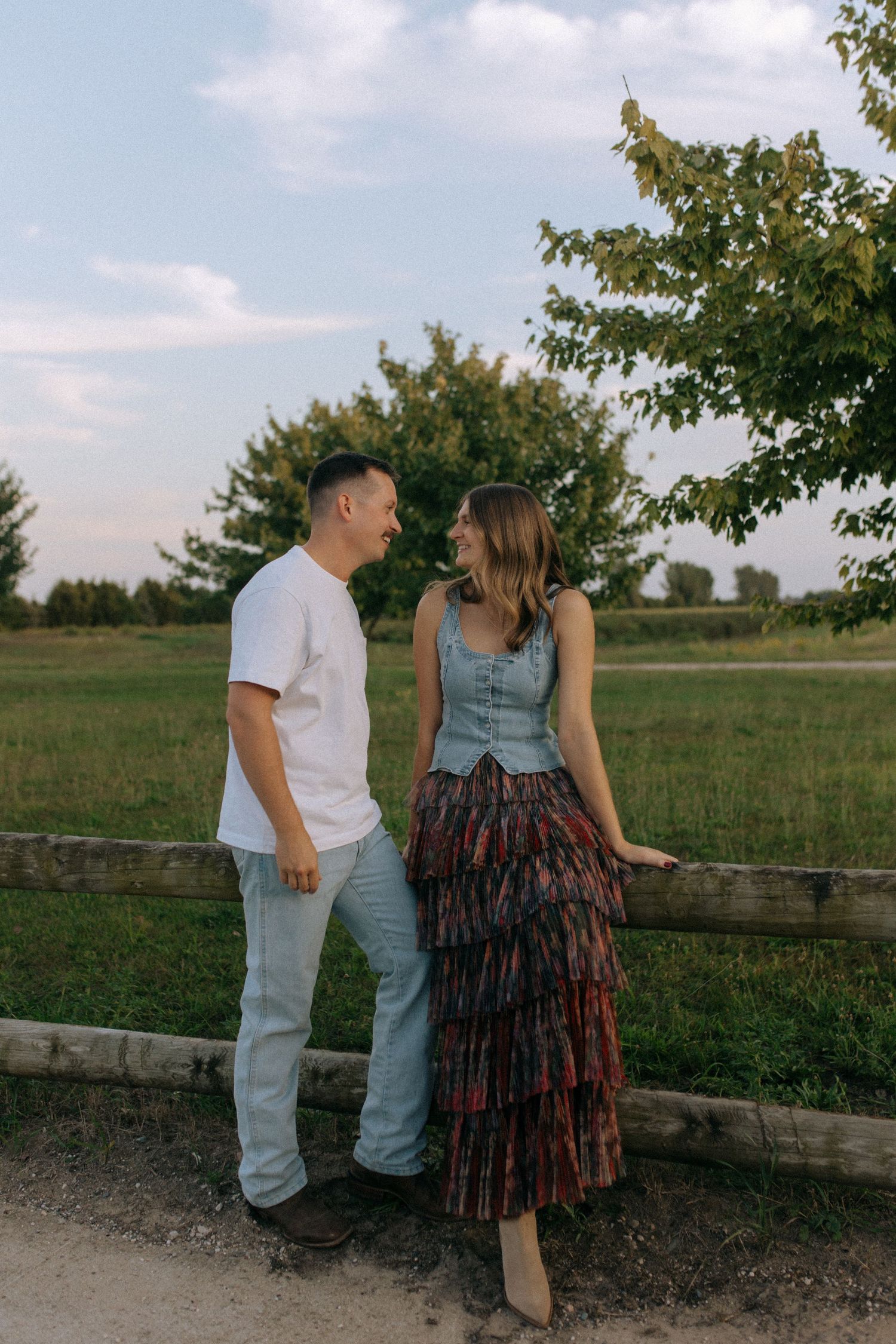 Two people pose casually against a wooden fence in a rural setting during daytime.