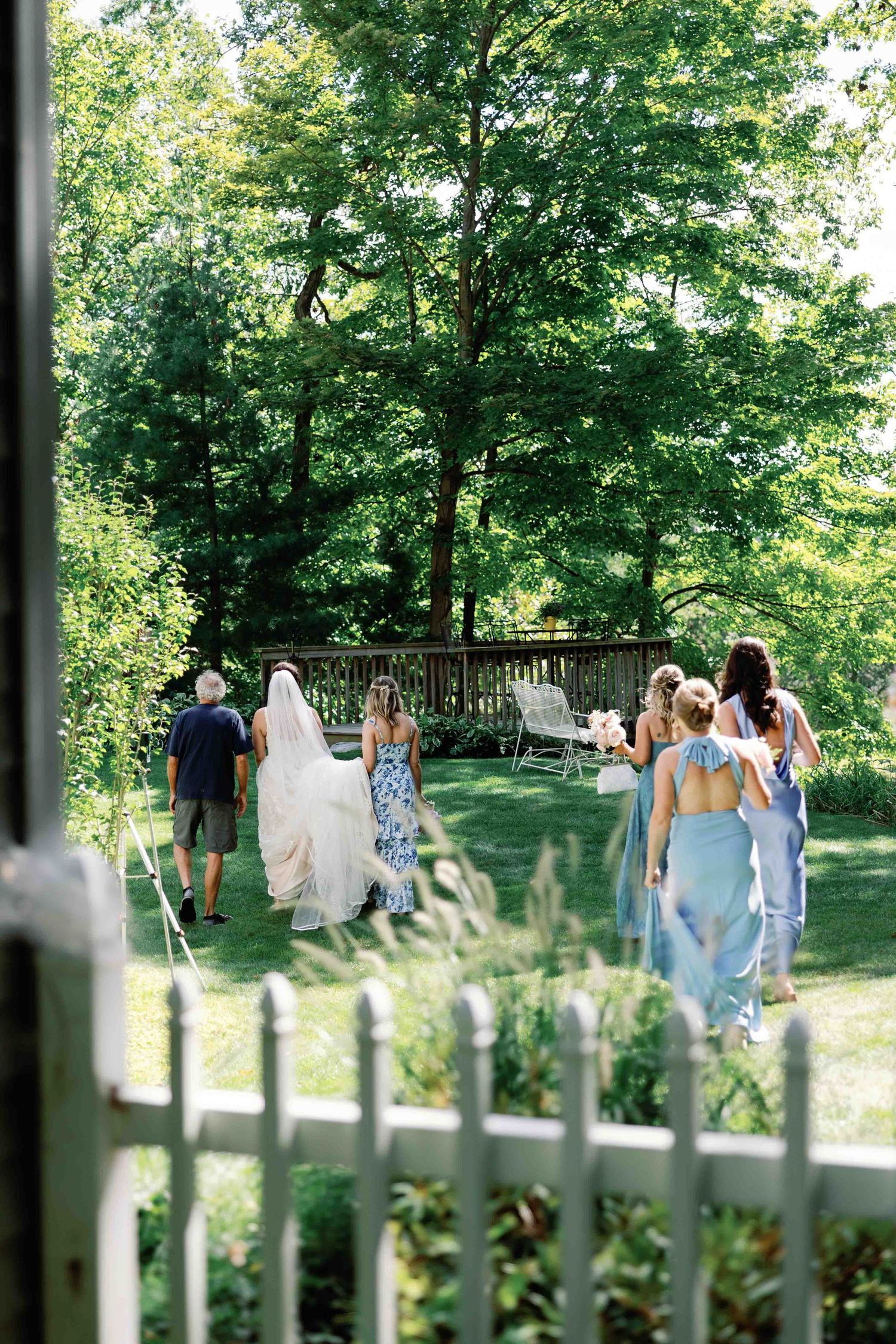 Wedding party walking through garden path viewed through decorative white fence.