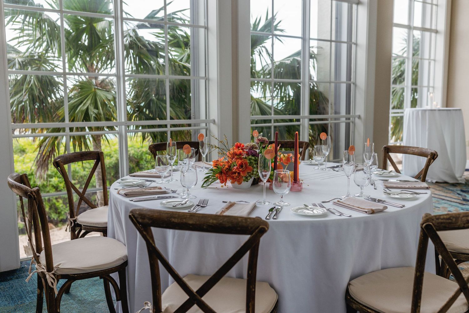 Elegant wedding reception tables set with white linens and palm trees visible through large windows in a bright venue.