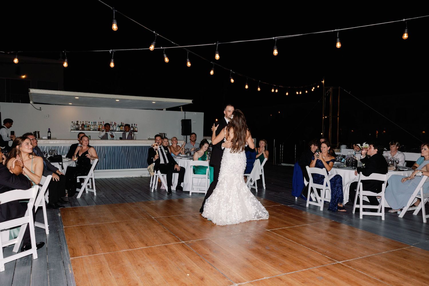 A bride in white dress dances at reception with guests seated at tables with white chairs.