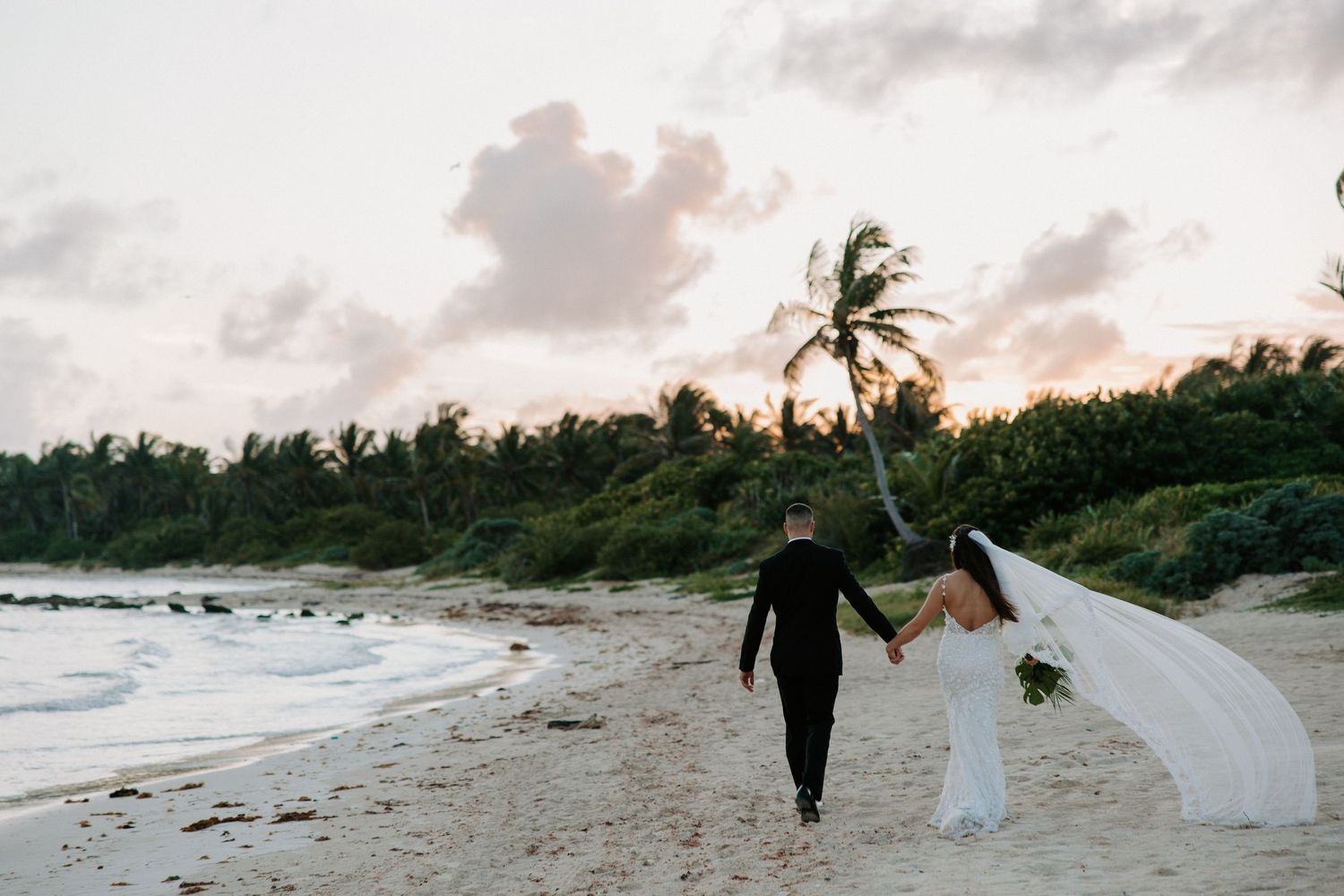 A couple walks hand in hand along a beach at sunset with palm trees in the background.