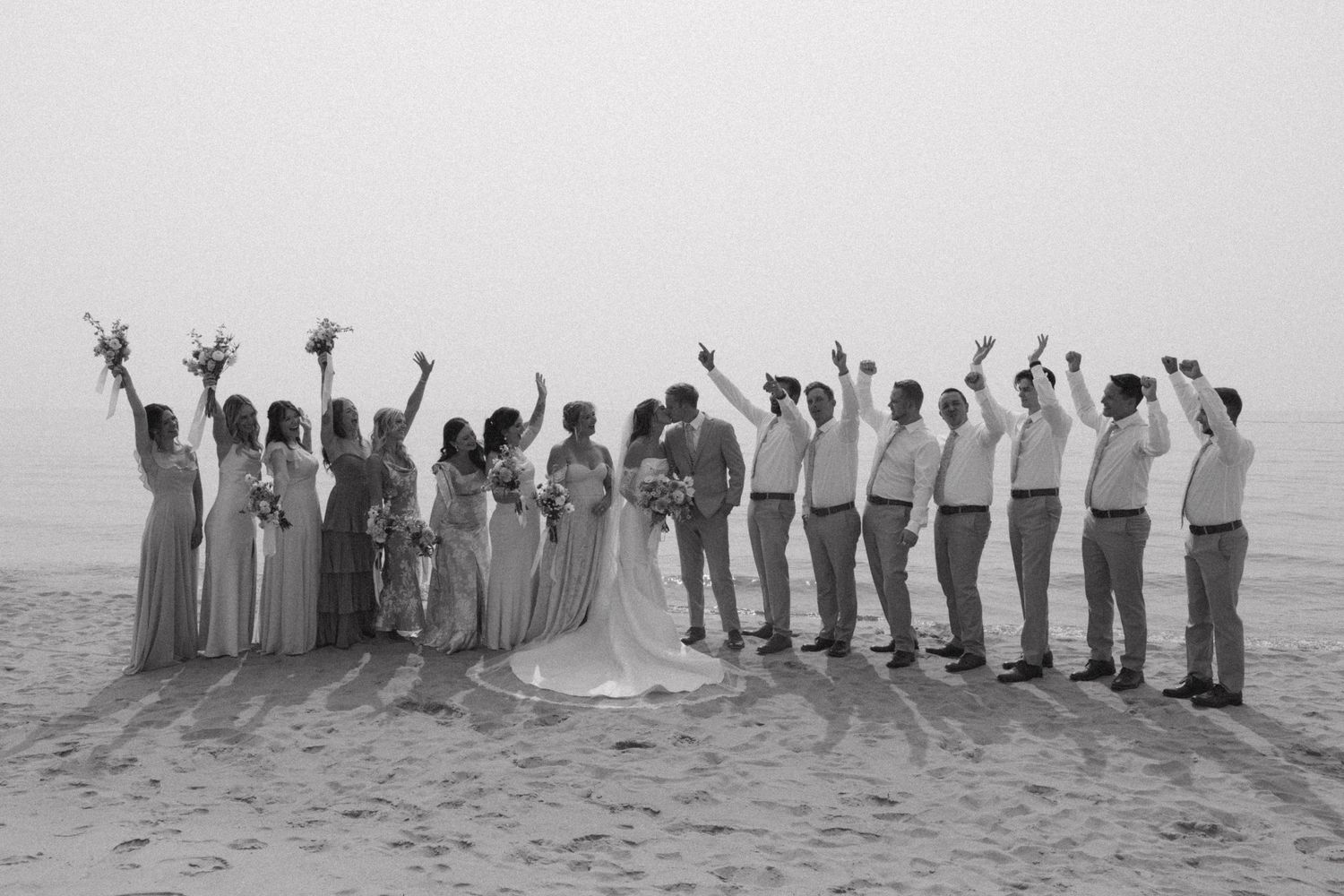 Wedding party lined up on a beach at sunset in shades of blue and gray attire.
