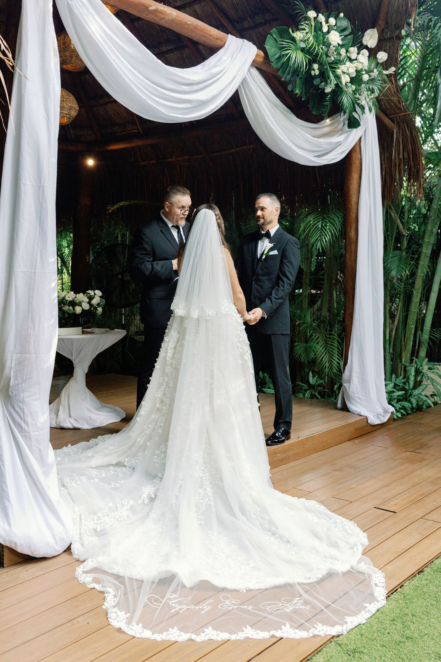 Elegant wedding ceremony under white draped fabric with trailing bridal gown on wooden deck.