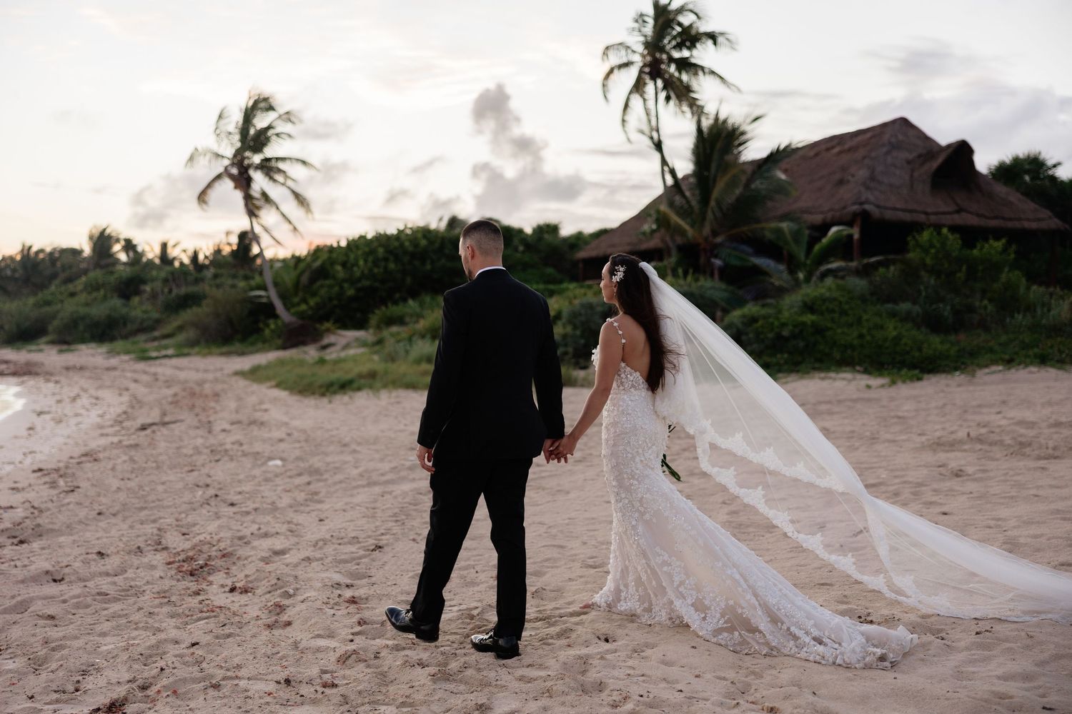 A couple stands on a sandy beach with palm trees and thatched roof buildings in background.
