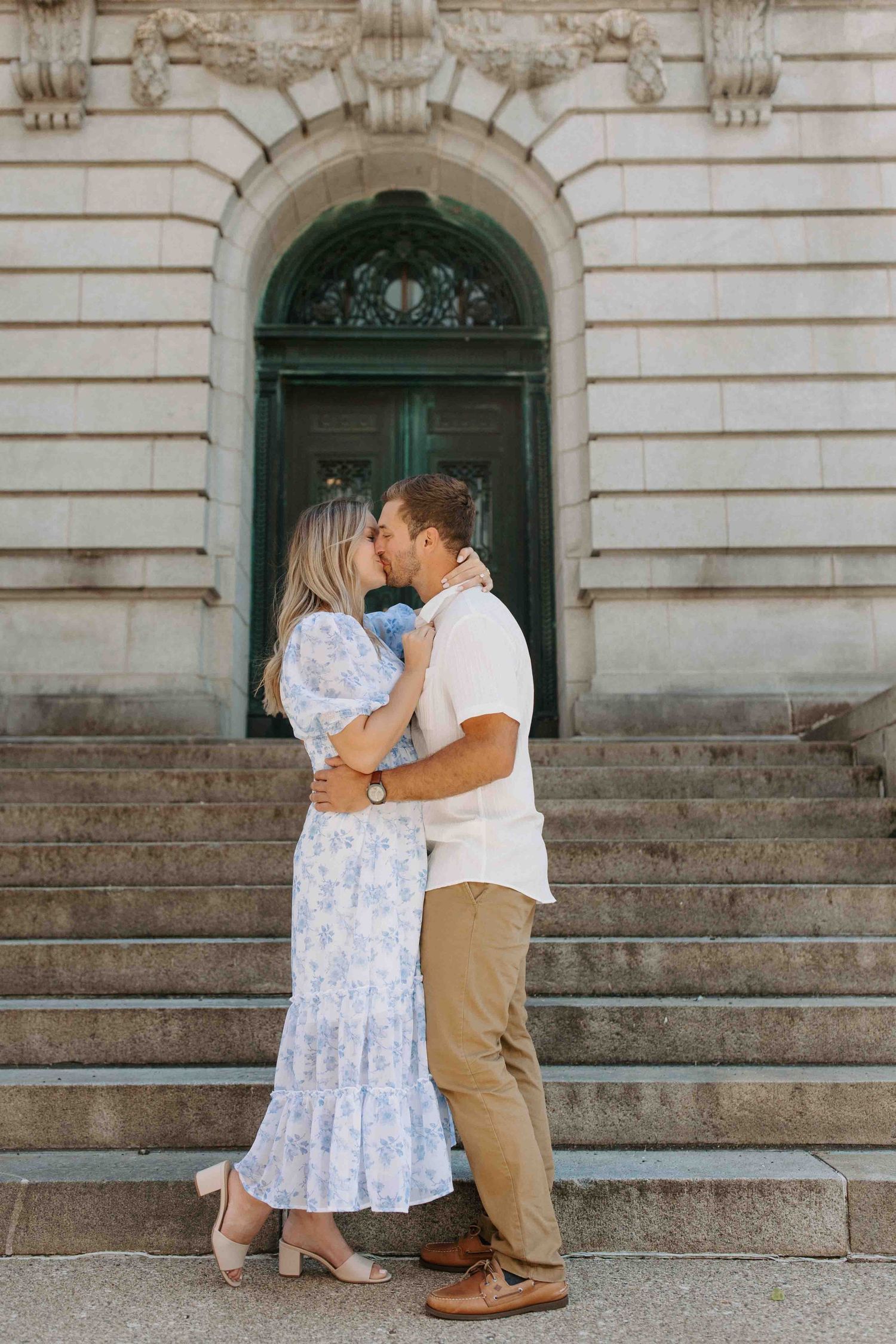 A couple embraces on grand stone steps of a classical building with ornate architecture.