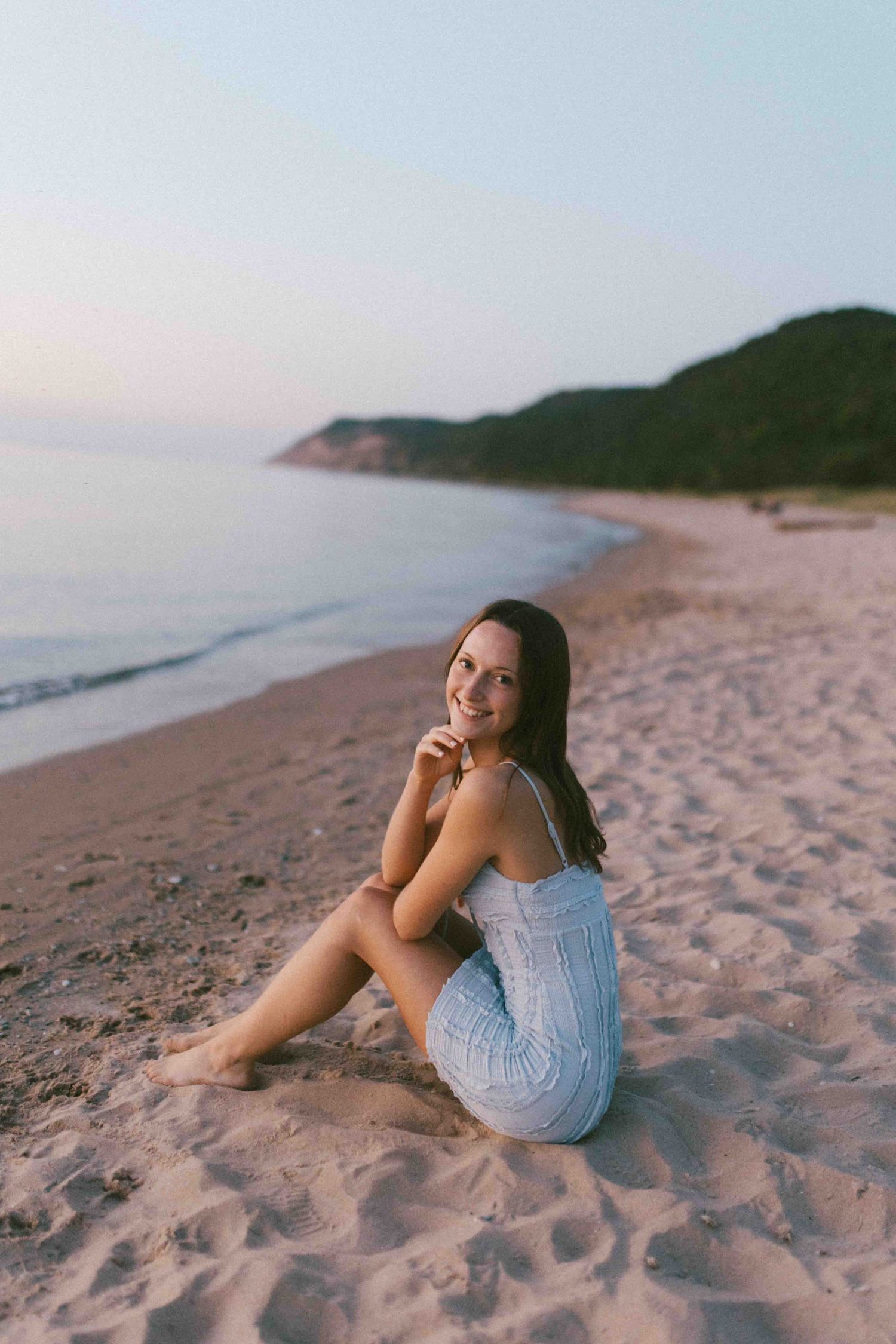 Person in blue dress relaxes on sandy beach at sunset with coastal cliffs in background.