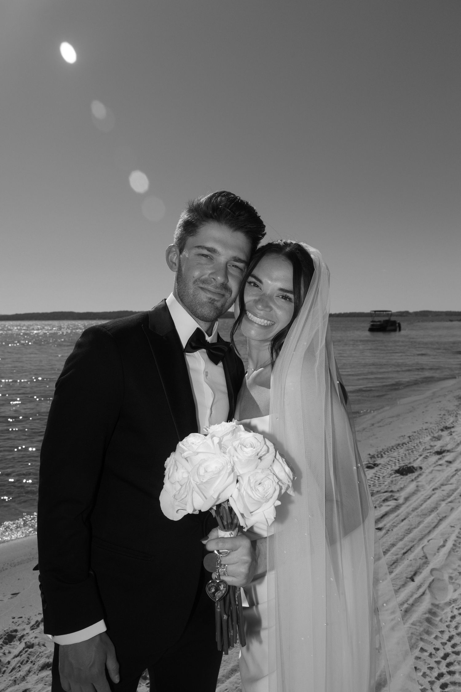 Newlyweds share an intimate moment on a sunny beach with white roses and ocean in the background.