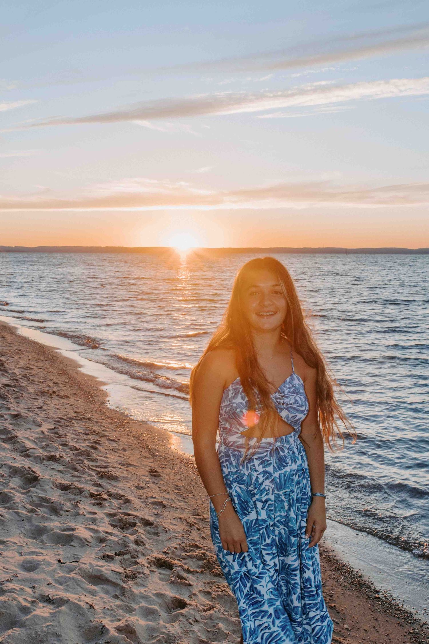 Person in blue floral sundress poses on sandy beach during golden hour sunset.
