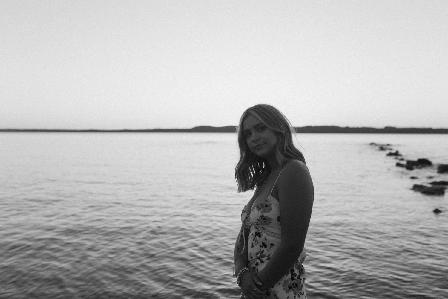 A figure stands by the water's edge during sunset in black and white photography.