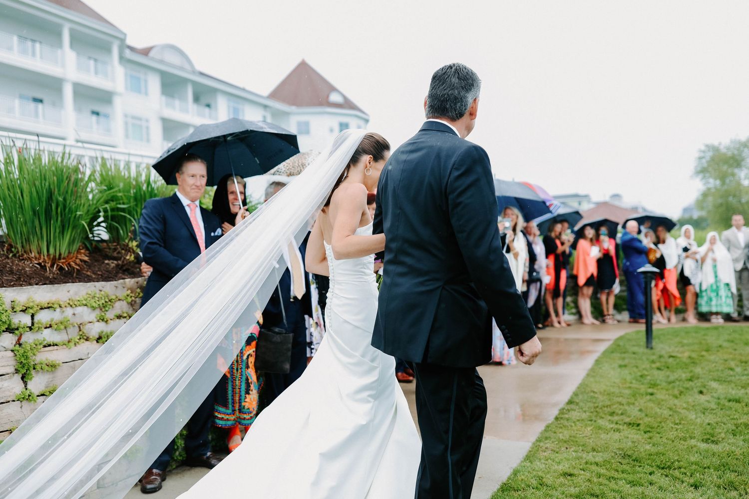 Wedding guests gather under umbrellas during an outdoor ceremony by the water.